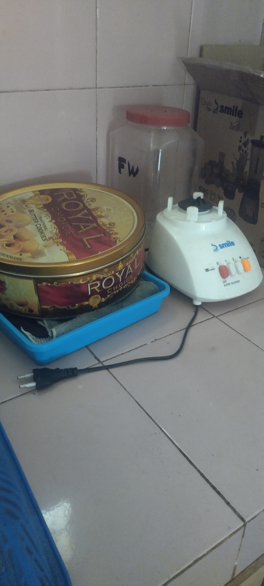 In a kitchen, on a tiled countertop, a round tin of Royal Choice butter cookies sits on a blue plastic tray. Next to it, a white Smile Super Blender is plugged in with a black cord and plug. Behind the blender, a clear plastic jar with a red lid is visible, with "FW" written on it. To the right, a cardboard box with the Smile logo is partially visible. The kitchen has tiled walls and floor.