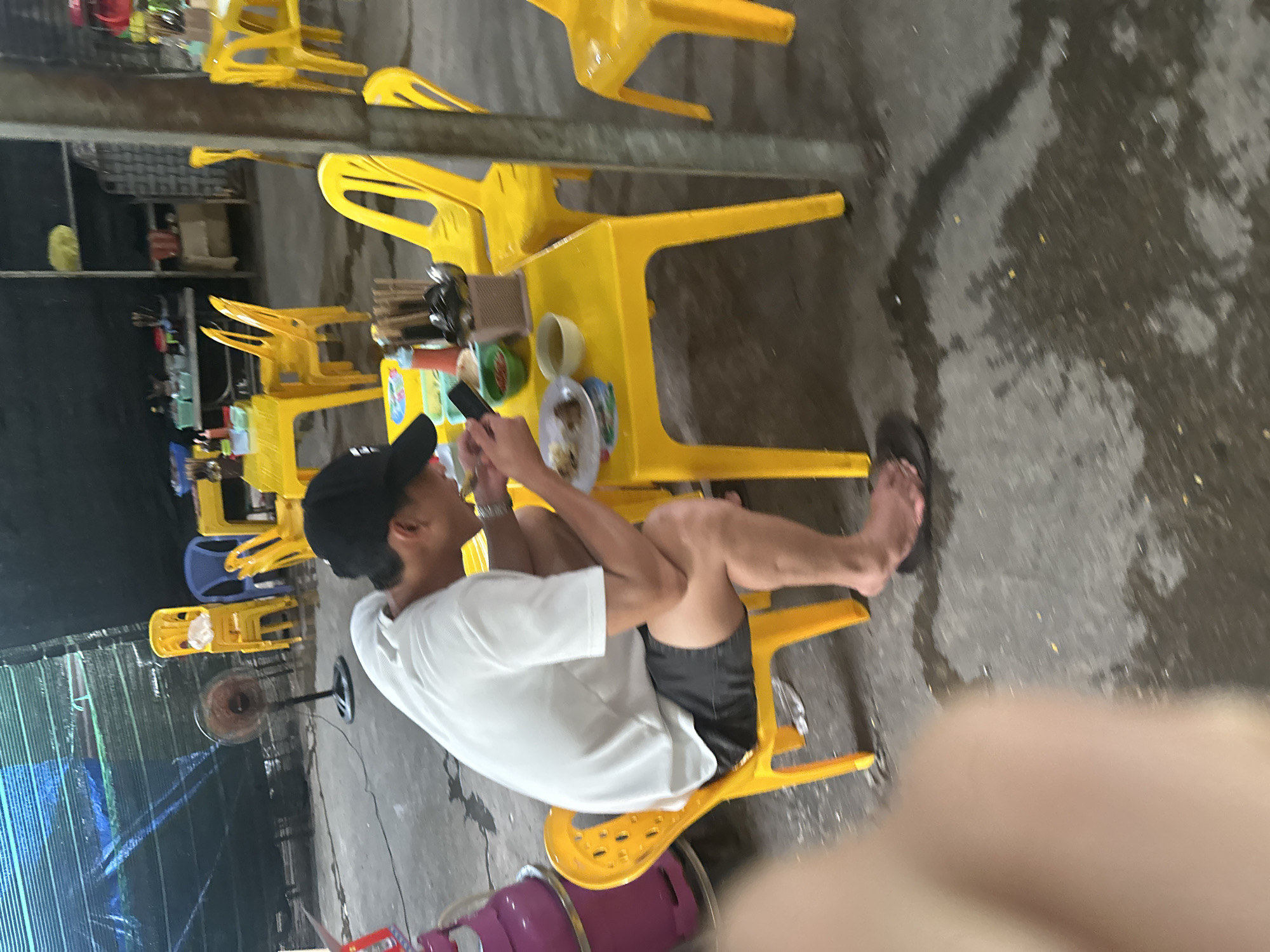 The image captures a casual eating scene, likely at a street food stall or a small eatery in Nha Trang Ward, Vietnam, as indicated by the context. The main subject is a man seated sideways on a yellow plastic stool, facing right in the upright orientation of the image. He is wearing a white t-shirt, black shorts, a black baseball cap, and flip-flops on his bare feet. He is holding a smartphone with both hands, engrossed in looking at its screen.
In front of him, on a yellow plastic table, rests a white plate containing what appears to be small food items or snacks, along with a white cup and small green containers. The dining area is characterized by numerous bright yellow plastic chairs and tables, a common sight in such establishments. In the background, another blue plastic chair is visible among stacks of yellow chairs. A standing fan is also positioned nearby, suggesting a warm climate. Further back, shelves with various items and a green mesh or net structure can be seen.
The concrete floor beneath the tables is damp and appears somewhat uneven, with visible puddles or wet patches, which could be from recent cleaning or rain. Part of a purple gas cylinder is visible in the bottom left foreground. The lighting suggests it could be late afternoon or evening, typical hours for casual dining. No discernible text is visible on any objects. The overall atmosphere is relaxed and informal.