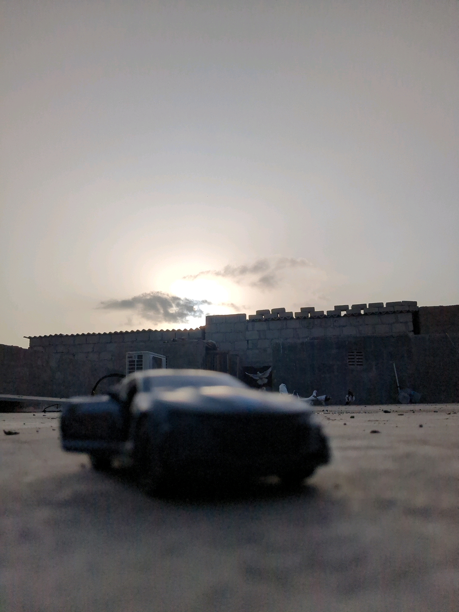 The image depicts a low-angle, close-up shot of a dark, blurry toy car in the foreground, positioned on a gritty, concrete surface. Behind the car, a weathered, concrete wall stretches across the mid-ground, topped with a decorative crenellated parapet. Above the wall, the sky is overcast with a soft, diffused sunlight breaking through, suggesting it is either sunrise or sunset. In the background, on top of the wall, a white air conditioning unit is visible. Further to the right, on the ground behind the wall, several objects are scattered, including what appear to be white birds or doves, and some debris. The overall scene evokes a sense of an urban or semi-urban environment, possibly on a rooftop or open ground. The specific location in the city of Karachi Division, Pakistan, is not explicitly identifiable from the visual elements alone.