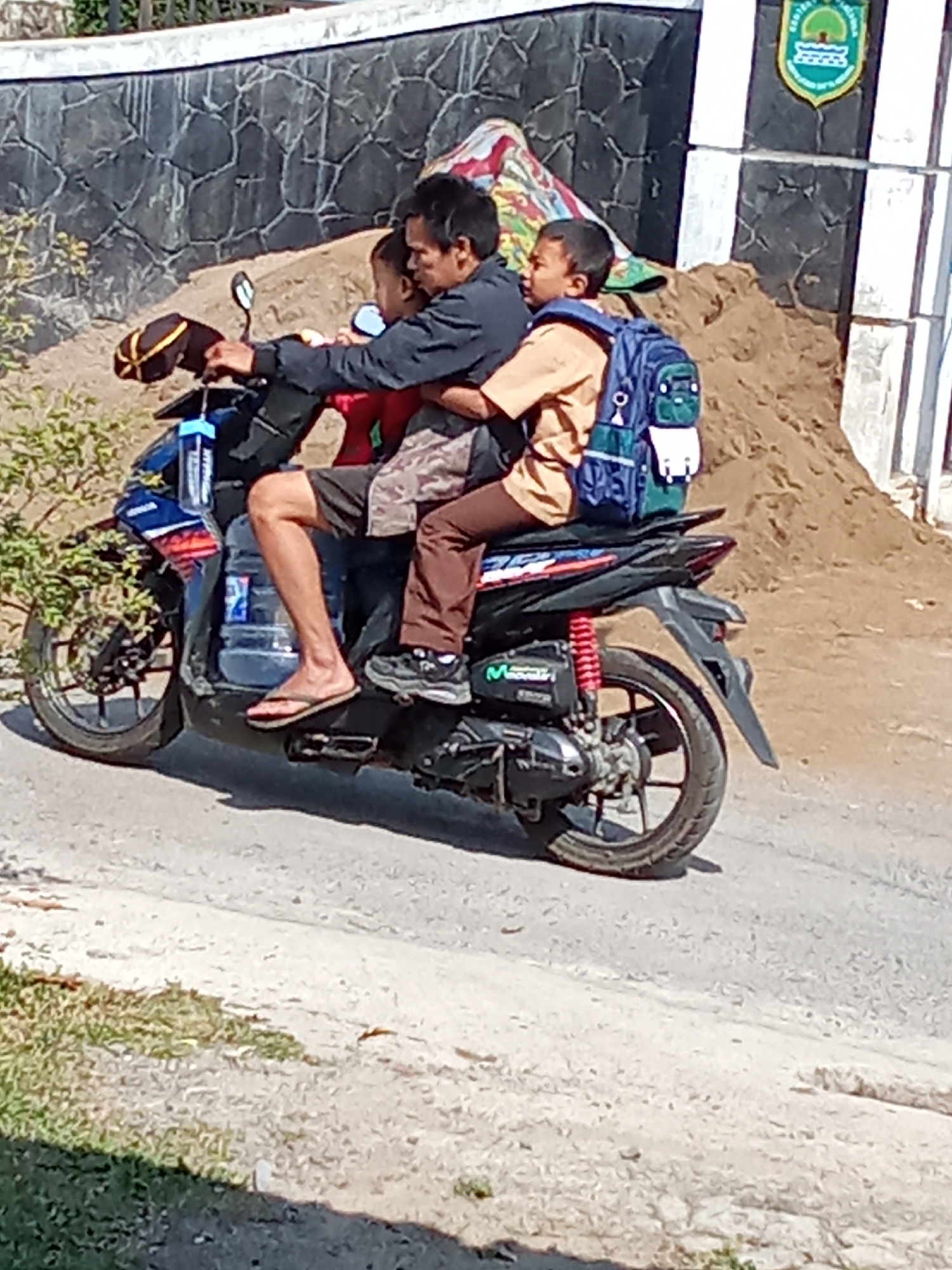 A father is riding a motorcycle with his two sons in Subang, Indonesia. The father is wearing a dark jacket and is in the front, with one son behind him, also wearing a dark shirt. The older son is in the very back, wearing a light brown school uniform shirt and a blue backpack. The motorcycle is dark blue and black, with a large water bottle attached to the front. The father is wearing flip-flops, and the older son is wearing athletic shoes. They are on a paved road with a slight incline, with a stone wall and a pile of dirt behind them. The wall has a crest with green and white colors at the top. The area appears to be rural or on the outskirts of a town.