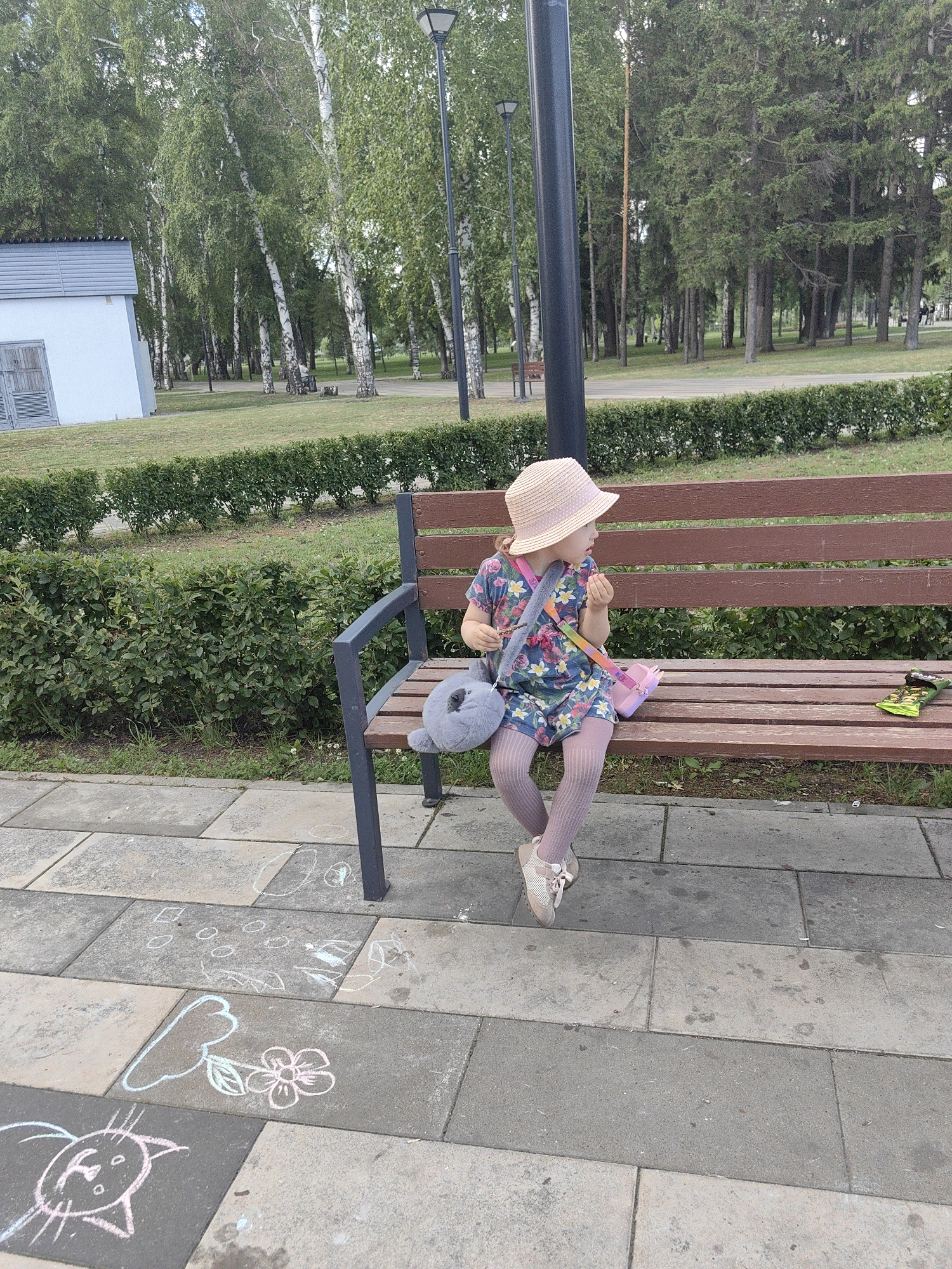 A young girl wearing a straw hat, a floral dress, and pink tights sits on a park bench. She has a gray plush toy in her lap and a pink bag slung over her shoulder. To her right on the bench is a green snack wrapper. In front of the bench on the paved walkway, chalk drawings of a cat and a flower are visible. The park appears to be in a city, possibly Ufa, Russia, based on the context of previous images from the same series. The background shows a line of birch trees and other greenery, with a lamppost and a small building in the distance. The lighting suggests it is daytime.