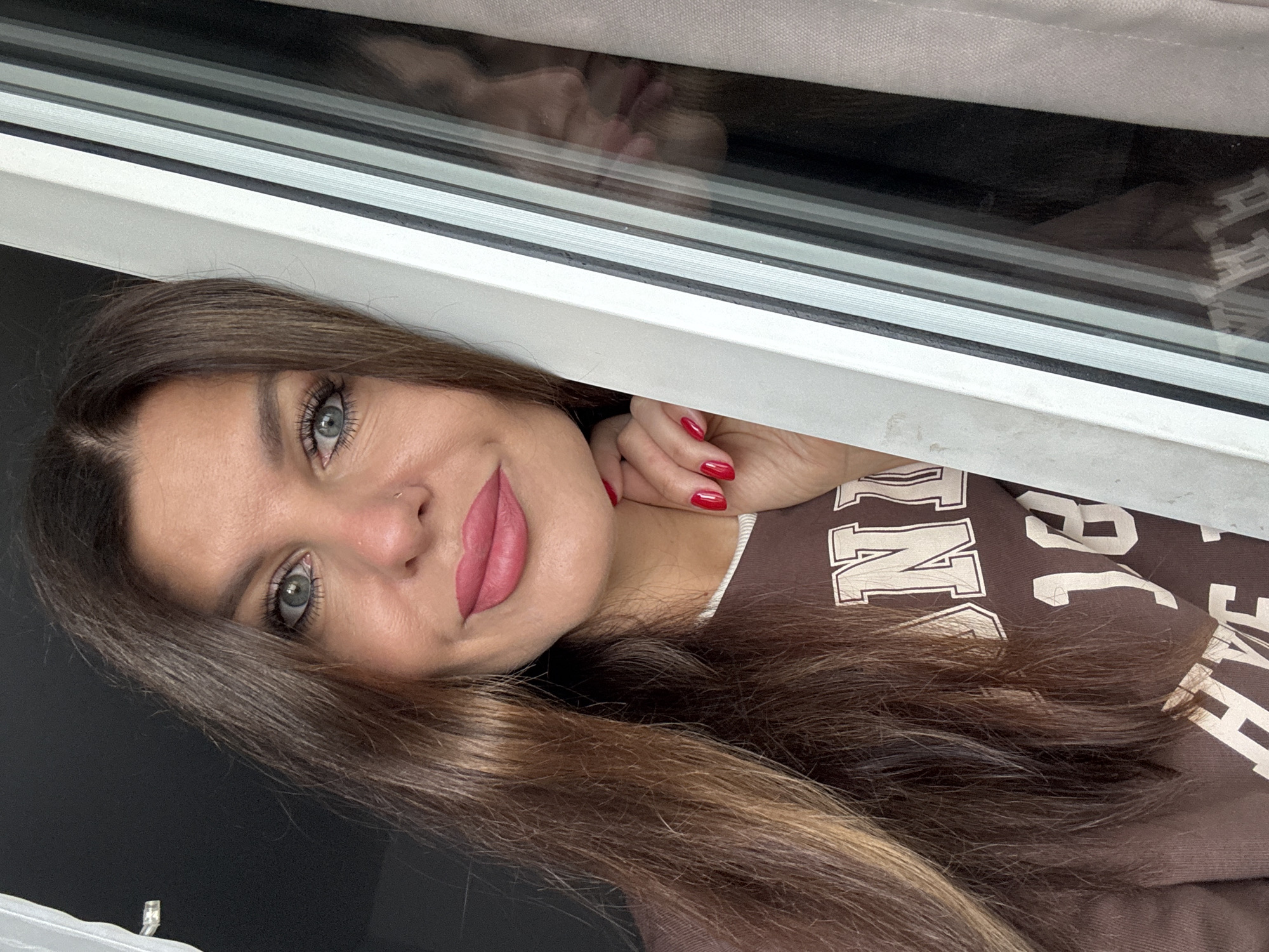 A medium shot captures a woman with long, brown hair, her face angled to the right. She is wearing a brown long-sleeved shirt with white lettering, and her left hand, with bright red nails, rests on her chin. Her eyes are blue, and she is wearing pink lipstick. The background features a window frame with white and black vertical accents, and a white curtain. A reflection of the woman is visible in the windowpane, her face also turned to the right. The scene appears to be indoors.