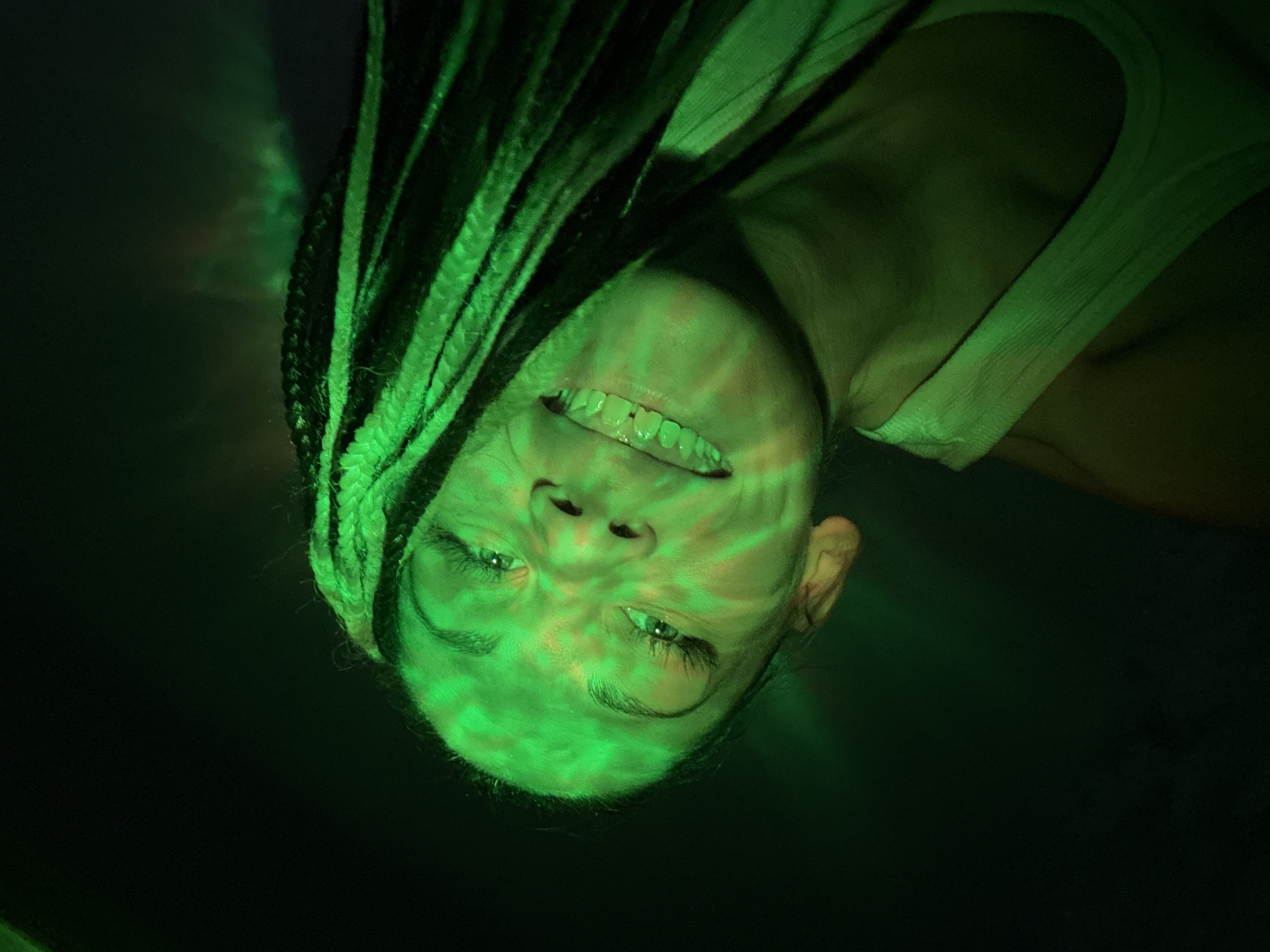 The image presents a close-up portrait of a person, likely female, captured in a dark environment illuminated by vibrant green light. The subject's head is angled, giving the impression of an upside-down or tilted perspective, with their face looking directly towards the camera. They have long, dark braided hair (dreadlocks or similar style) that frames their face, with some braids dramatically highlighted by the intense green light.
The person is wearing a light-colored, possibly white, tank top or camisole, part of which is visible on their right shoulder. Their face is animated with a wide, open-mouthed smile, revealing their teeth and suggesting an emotion of joy, laughter, or amusement. Their eyes are open and focused, and appear to be a light color, reflecting the green illumination. Dynamic, patterned green light is projected across their face and upper body, creating swirling lines and shapes that enhance the energetic atmosphere.
The background is predominantly dark, almost black, serving to emphasize the bright green light and the illuminated subject. The scene strongly suggests an indoor setting, likely at night, such as a nightclub, concert, party, or a creative photo shoot utilizing special lighting effects. There are no discernible objects other than the person and their clothing, nor is any visible text present. The overall mood is lively and engaging. Given the location context, this event takes place in Rusanovo, Russia.