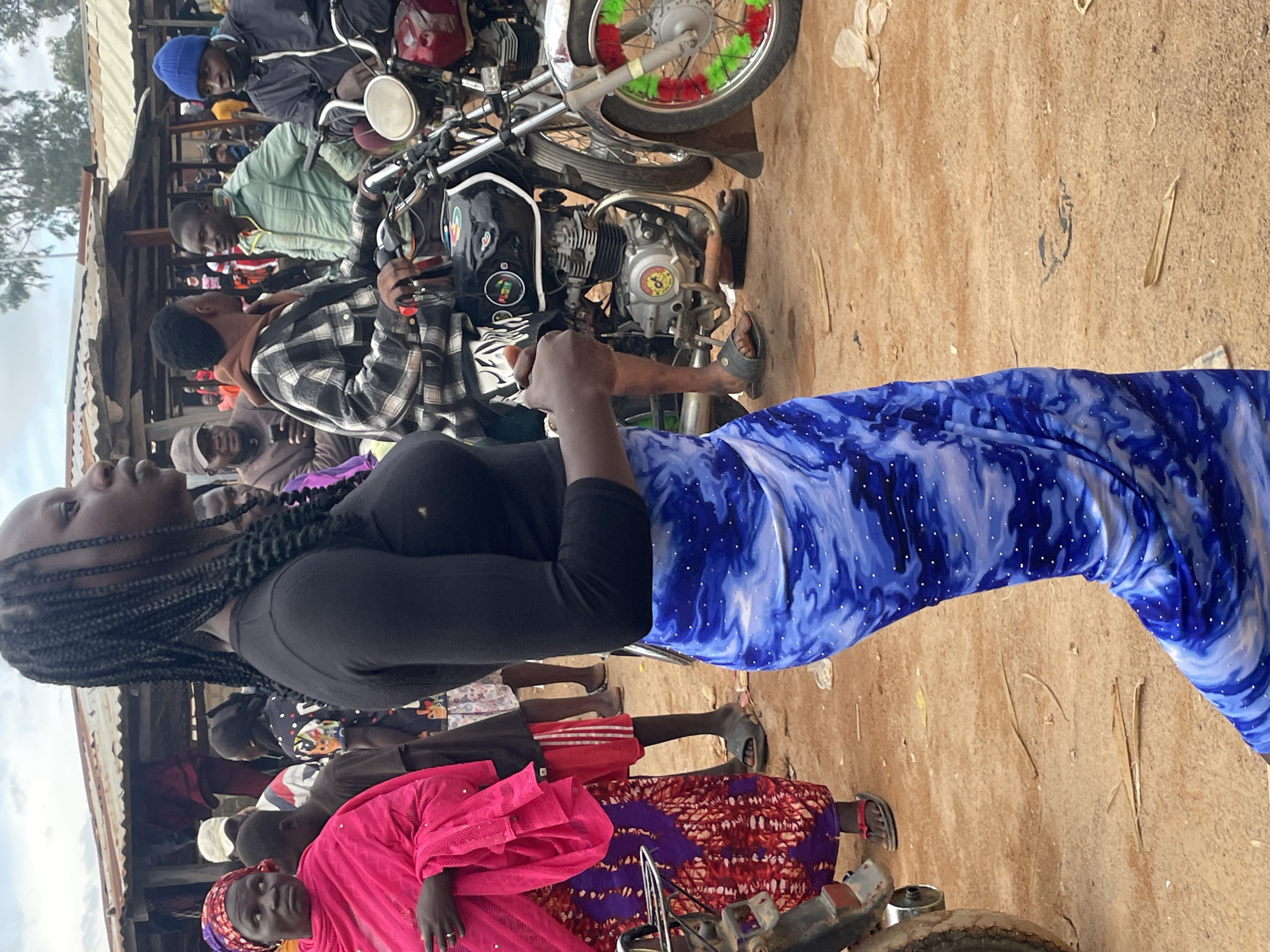 The image depicts a bustling outdoor scene in Vom, Nigeria, likely a marketplace or a busy street. In the foreground, a young woman with braided hair, wearing a black long-sleeved shirt and a flowing blue and white patterned skirt adorned with small white dots, is the primary subject. Her gaze is directed to the right, and she is positioned slightly off-center. To her left, another woman is visible, wearing a vibrant pink headscarf and a patterned purple and red skirt.
Behind the main subjects, several people are present, some interacting with motorcycles. One man in a plaid shirt is seated on a black motorcycle, looking towards the woman in the foreground. Another man to his right wears a green jacket and appears to be talking. A third man, wearing a blue beanie and a dark jacket, is also near a motorcycle, with a portion of its front wheel visible, decorated with red and green accents. The environment is characterized by dusty, reddish-brown ground, typical of outdoor settings in many Nigerian cities. Several stalls or makeshift structures with corrugated metal roofs are visible in the background, suggesting a commercial area. The overall impression is one of everyday life unfolding in a lively, perhaps somewhat crowded, urban environment.
