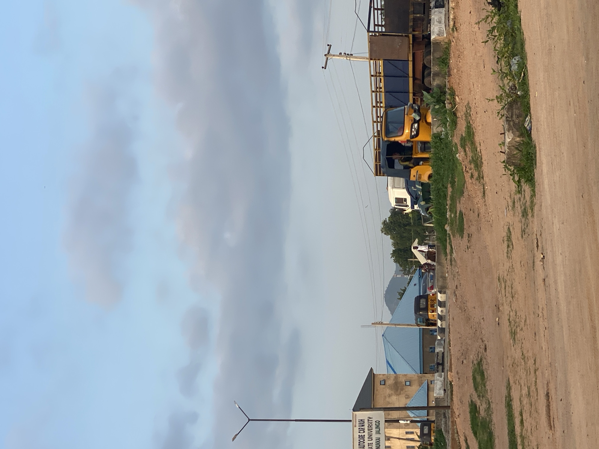 The image shows a street scene in Jalingo, Nigeria, with a focus on a road intersection. In the foreground, there is a dirt road with sparse vegetation. To the left, a signpost reads "AUTOCARE CAR WASH" and "STATE UNIVERSITY, NUMKAI JALINGO." Behind the signpost, there is a building with a beige facade and blue tiled roofs.
In the middle ground, a yellow auto-rickshaw is parked at the intersection, and another one is visible further down the road. A large truck with a blue trailer is parked next to the auto-rickshaw. There are also a few people standing around, and a white vehicle is parked behind the yellow auto-rickshaw. The background features more buildings with blue roofs, trees, and a hill in the distance. The sky is partly cloudy. The scene appears to be during the day, possibly late afternoon given the lighting.