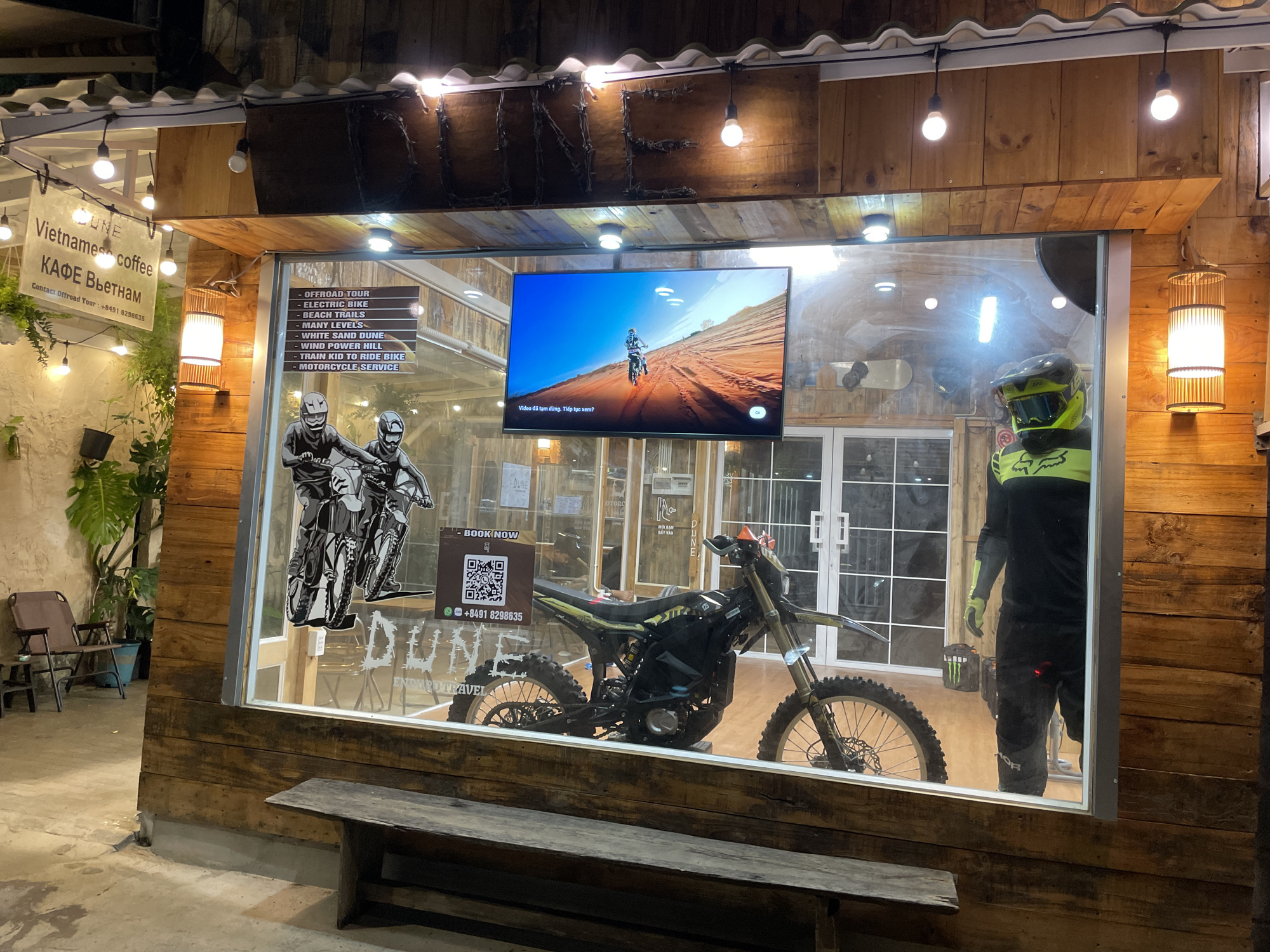 Here is a description of the image:
The image depicts the storefront of "DUNE Enduro Travel" in Phu Thuy Ward, Vietnam. A window displays promotional materials for off-road tours and electric bikes. A television screen shows an off-road motorcycle video. A man in motorcycle gear stands to the right. A black and yellow dirt bike is prominently displayed. A sign advertises "Vietnamese Coffee" next to the main storefront. A wooden bench sits in front of the store.