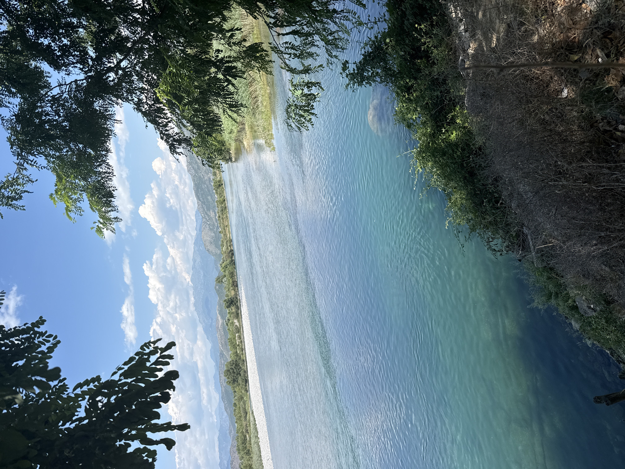 The image depicts a serene natural scene with a wide river flowing through a valley. The water is a clear, vibrant blue, with gentle ripples on its surface. The riverbanks are lined with lush green vegetation and reeds, with a pathway of white stones running along one side. In the distance, majestic mountains rise, their peaks softened by a hazy, possibly summer, atmosphere. The sky above is a brilliant blue, dotted with fluffy white clouds, suggesting a pleasant, sunny day.
Framing the scene from the top and bottom left are the dark green leaves of trees, their branches creating a natural border and adding depth to the composition. The sunlight filters through the leaves, casting dappled shadows. On the right bank, the terrain becomes more rugged, with dry brush and scattered debris suggesting a less cultivated area. The overall mood is tranquil and picturesque, evoking a sense of peace and natural beauty. There is no visible text or discernible human activity within the frame.