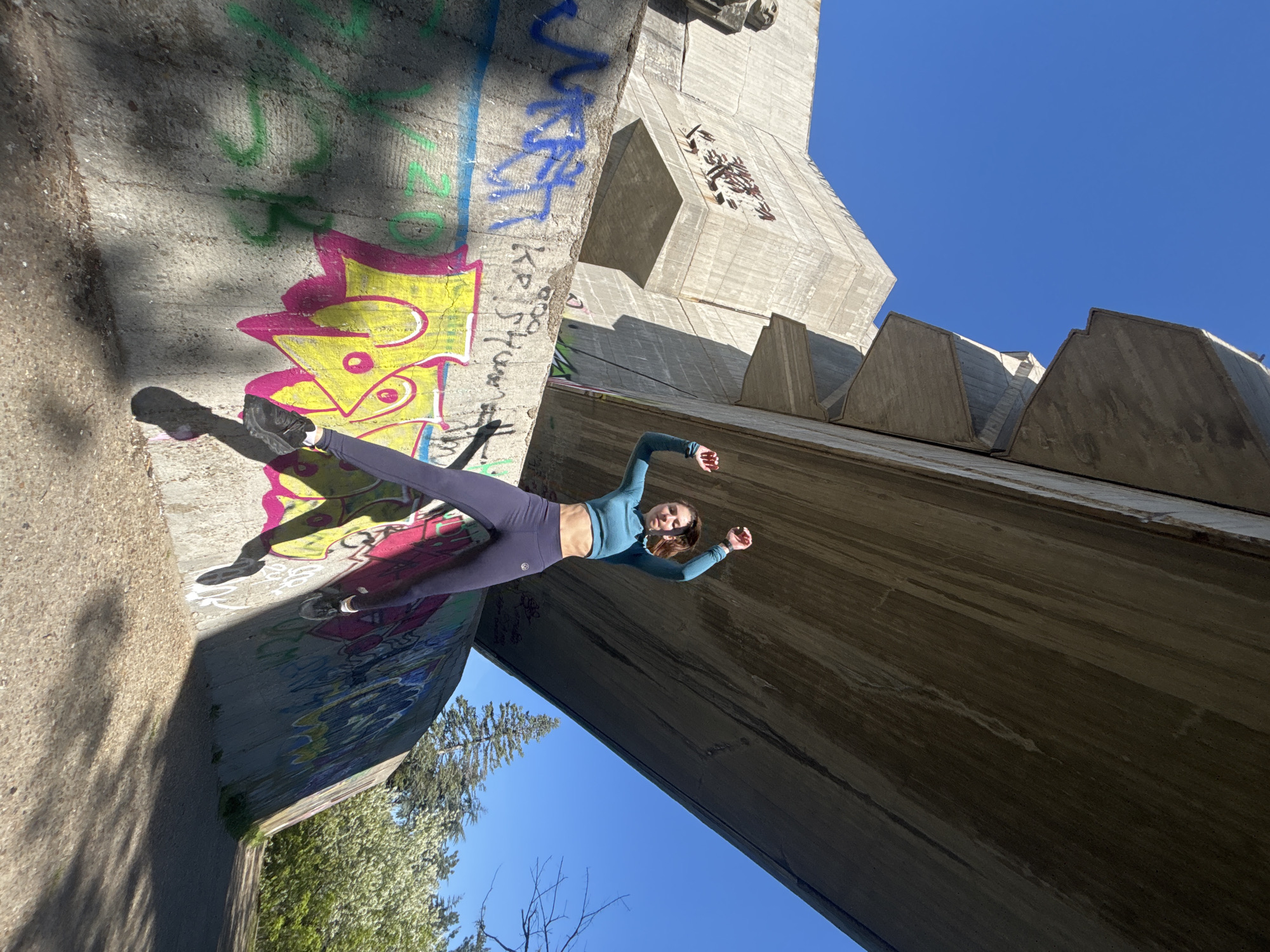 A young woman in athletic attire (blue crop top and purple leggings) is positioned between two concrete structures with graffiti on one side. The environment includes trees and a clear blue sky, suggesting an outdoor location. The image is taken from a low angle, looking up at the structures and the woman. Location is Unknown, Unknown.