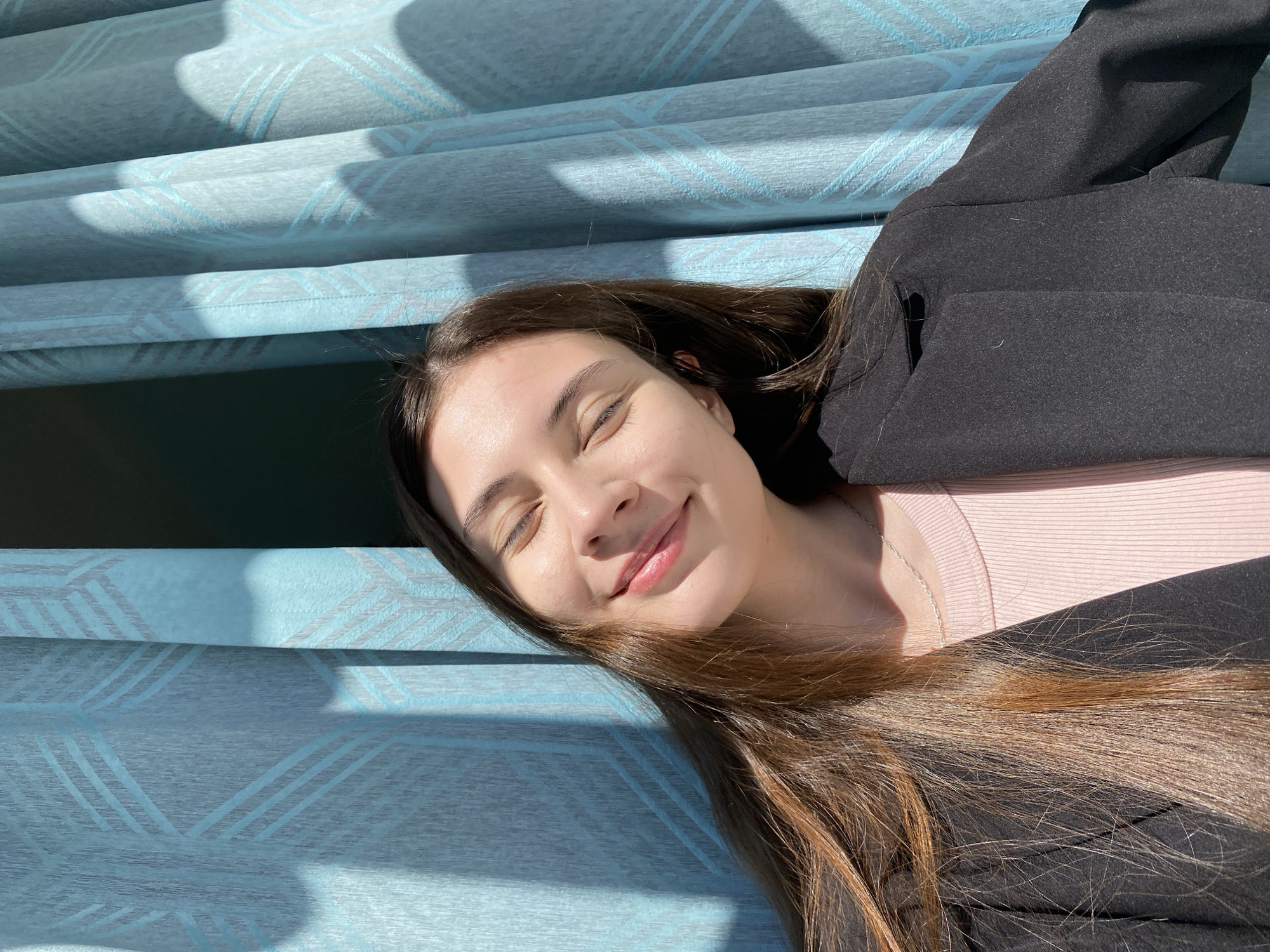 A young woman with long, brown hair, wearing a light pink top and a black blazer, is lying on a light blue surface with a geometric pattern, possibly a blanket or fabric. Her eyes are closed and she is smiling. The scene is bathed in sunlight, creating shadows on the surface. No information in the image indicates a specific location in Votkinsk, Russia, or any other city.