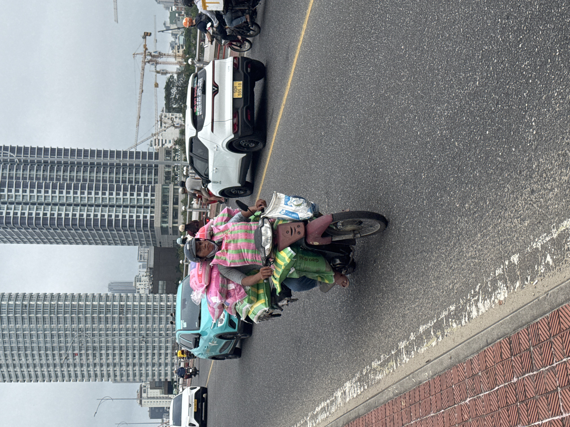 The image captures a street scene in Đà Nẵng, Vietnam, characterized by heavy traffic. A man on a motor scooter laden with pink and green bags and bundles navigates the road. He wears a grey helmet, a grey shirt, and blue jeans and rides barefoot. In the background, tall skyscrapers dominate the skyline under an overcast sky. The road is asphalt, marked with a yellow line separating lanes. Several cars and other motor scooters are visible in the background. The foreground shows a red brick pattern adjacent to the road. Construction cranes can be seen in the distance.