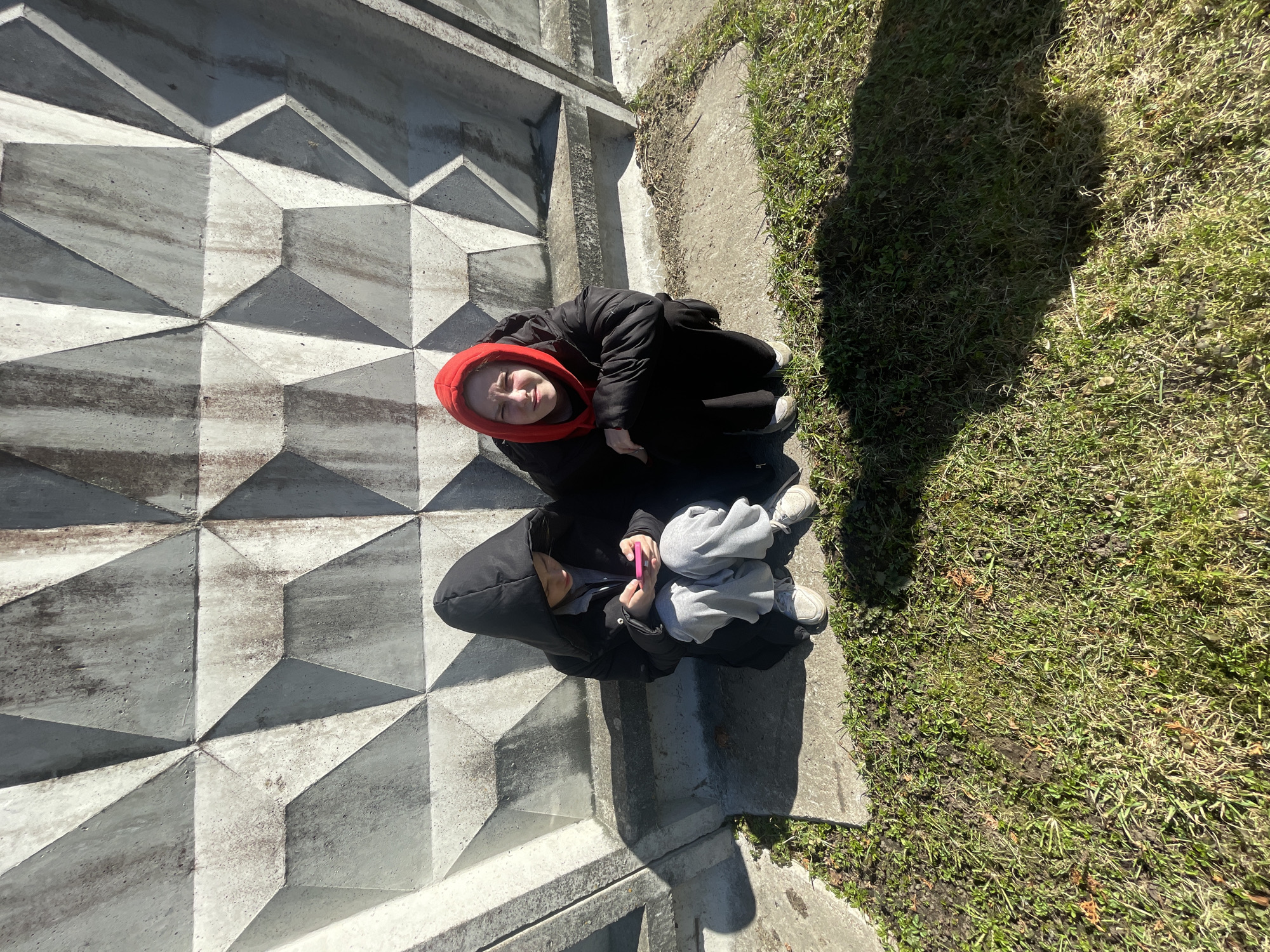 The image depicts a high-angle view of two individuals. One individual, wearing a black coat with a red hood, is looking upwards. The second individual, wearing a black hooded jacket, is seated below, holding a pink phone. The setting appears to be an urban area with concrete blocks and grass. It could be Saint Petersburg, Russia but more context or closer images would be needed to confirm.