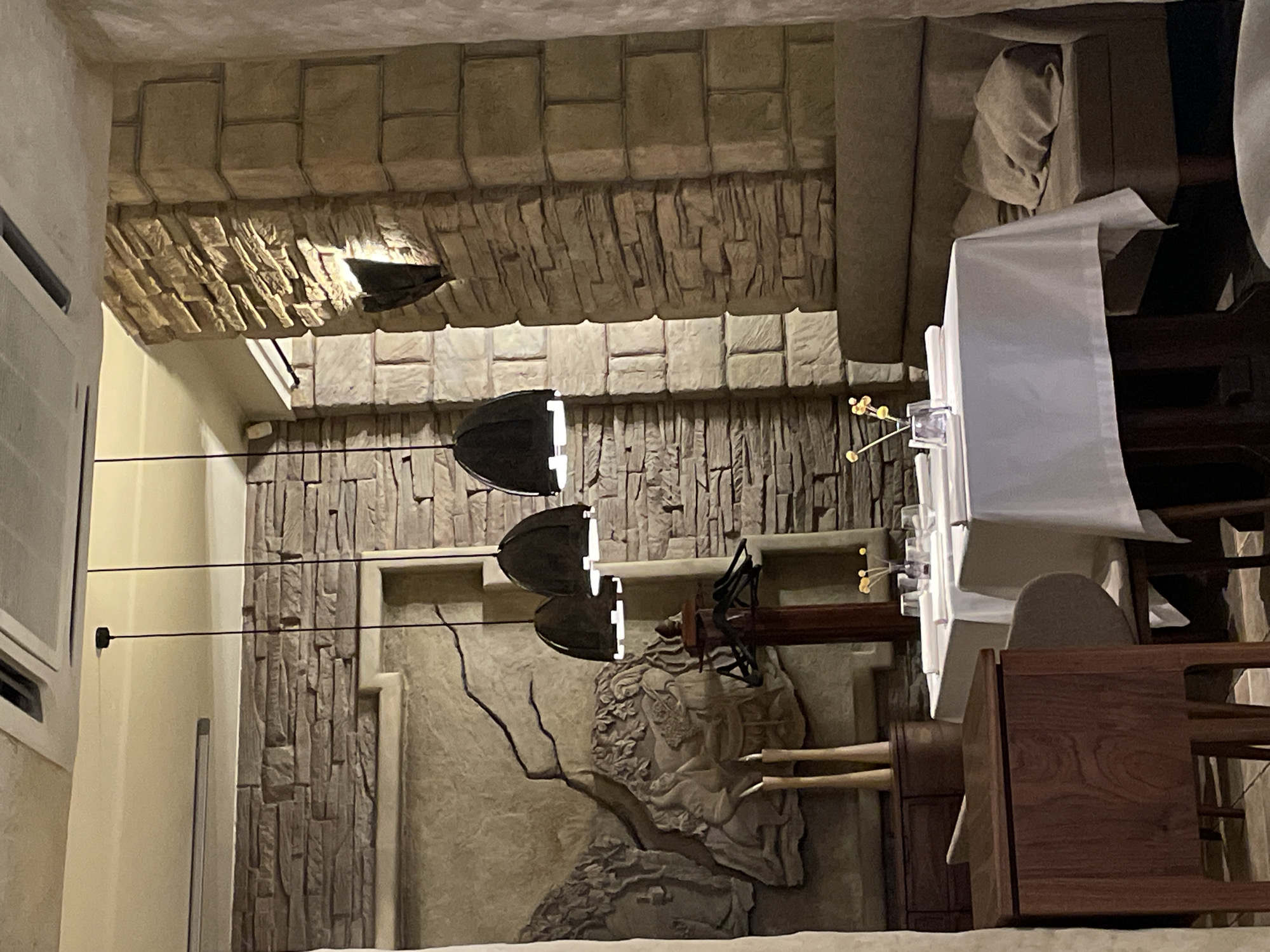 This image, when viewed in its correct upright orientation, depicts an inviting and sophisticated indoor dining area, likely a restaurant or an upscale private dining room. The walls are a prominent feature, adorned with textured stone cladding. The upper sections exhibit neatly laid, block-like stones, while the lower parts showcase more rugged, vertically oriented, rough-hewn stone elements, creating a blend of rustic and refined aesthetics.
Several dark, bell-shaped pendant light fixtures hang at different heights, casting a warm glow and providing the primary illumination for the space. Reflections of light are visible on their metallic surfaces. To the far left, a white air conditioning unit is partially visible on a light-colored wall.
In the foreground and to the right, dining tables are visible, covered with crisp white tablecloths and accompanied by chairs. One table holds a small vase with delicate yellow decorative elements. The absence of people suggests the image was captured either before or after dining hours.
A notable artistic detail is a large relief sculpture on the lower section of the wall. This intricate artwork appears to feature a figure, possibly mythological, intertwined with organic motifs resembling branches or vines, adding a classical and artistic touch to the decor. A dark wooden structure, potentially part of a serving cart or another piece of furniture, is partially visible near the tables.
The artificial lighting and the general atmosphere indicate an evening setting. The unique combination of natural stone, detailed relief art, and elegant table settings points to a location with a distinctive interior design. No visible text is present in the image.