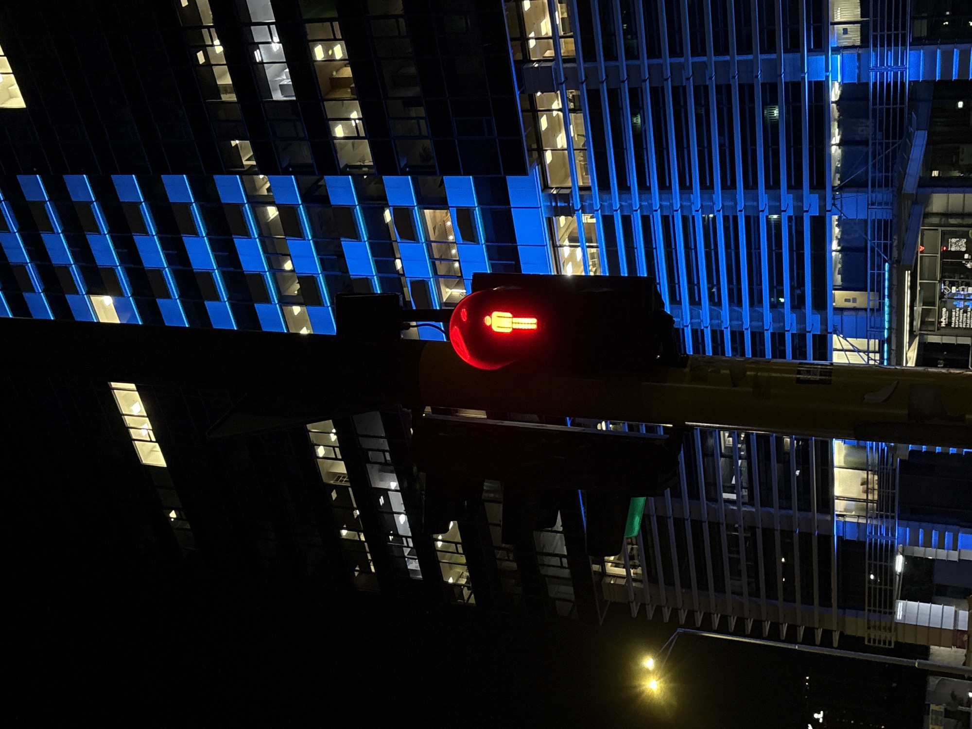This night photograph, taken in Sofia, Bulgaria, captures a striking urban scene dominated by artificial light and reflections. The image presents a view across a body of water, likely a canal or large puddle, reflecting the city's illuminated architecture.
In the center, a prominent pedestrian traffic light is vertically oriented, displaying an illuminated red "don't walk" symbol depicting a standing human figure. This light is affixed to a dark, horizontal structure that spans across the mid-ground of the composition.
To the right of the traffic signal, modern city buildings rise, showcasing distinct architectural lighting. One building features a dynamic checkerboard pattern of bright blue squares, while an adjacent structure is adorned with vertical blue light strips. Numerous windows within these buildings glow with warm yellow light, offering glimpses into interior spaces.
The left side of the image is filled with the clear, mirrored reflections of these urban structures and their vibrant lights in the water. These reflections create an almost symmetrical and abstract visual, repeating the blue, yellow, and dark tones of the actual buildings. Scattered yellow-orange lights, possibly from streetlights or distant windows, are visible in the lower left portion of this reflected area.
The scene conveys the serene yet vibrant atmosphere of a city at night, with the interplay of architectural lighting and its precise reflection being the primary visual focus. No human activity is depicted, and the only visible text is the iconic pedestrian symbol on the traffic light.
