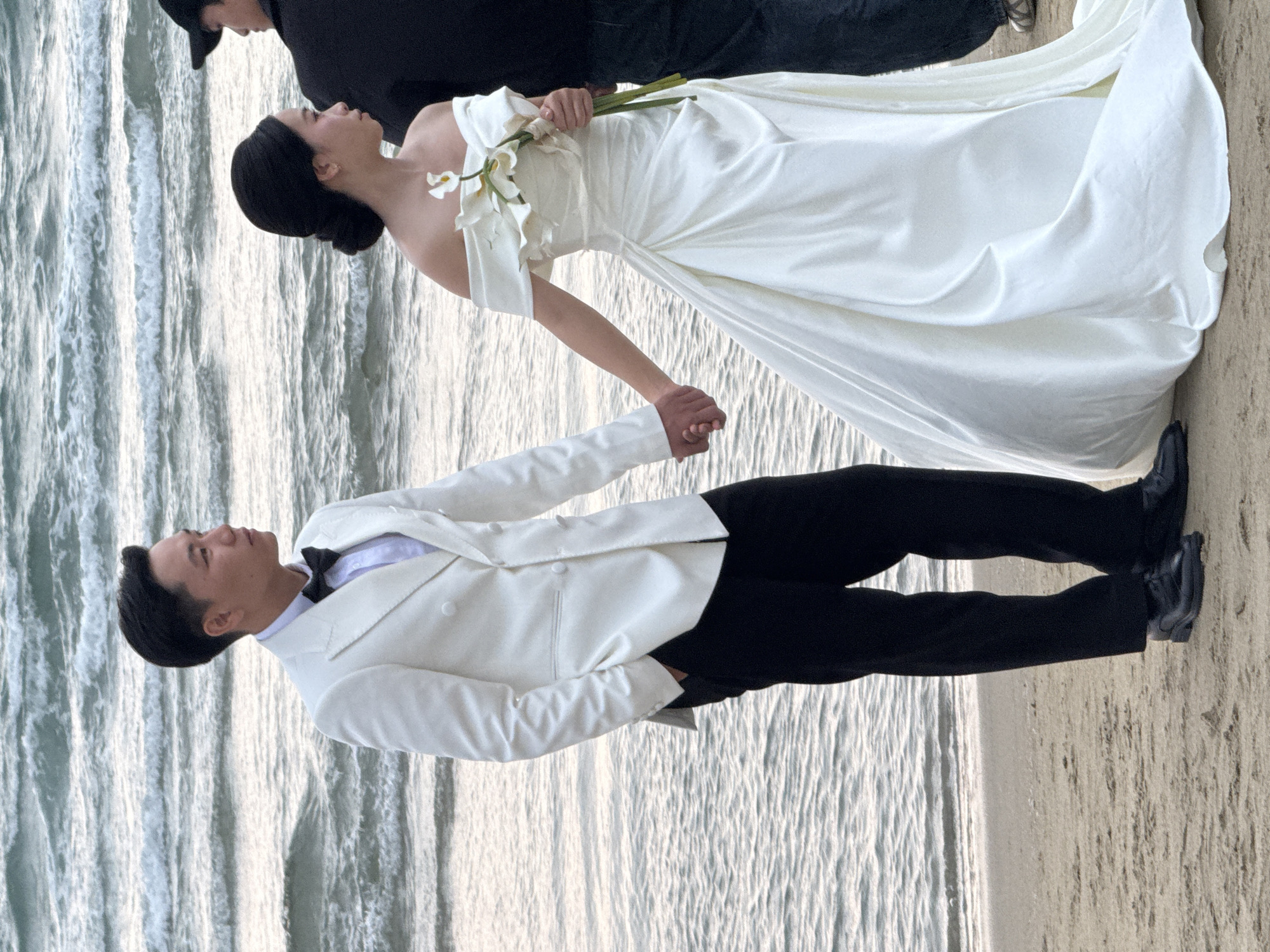 This image captures a wedding couple posing on a beach in Đà Nẵng, Vietnam. The bride, positioned on the left, wears an elegant, flowing white satin wedding gown with delicate off-the-shoulder sleeves. Her dark hair is styled in a neat bun, and she holds a bouquet of white calla lilies in her left hand. She looks upwards and slightly to her left, with a serene expression. Her right hand is clasped with the groom's left hand.
The groom, standing to the right of the bride, is dressed in a formal attire consisting of a white double-breasted suit jacket over a white shirt, a black bow tie, black trousers, and black dress shoes. He looks slightly to his right, away from the camera. A third individual, largely obscured in the background behind the bride, is seen wearing a black cap and black clothing.
The setting is a picturesque shoreline, with the gentle, rippling waves of the ocean occupying the left side of the frame, reflecting the bright daylight. The right side of the image reveals light-colored sand. The lighting suggests a clear day, possibly late afternoon, creating a soft and romantic atmosphere for what appears to be a wedding photoshoot. There is no visible text in the image.