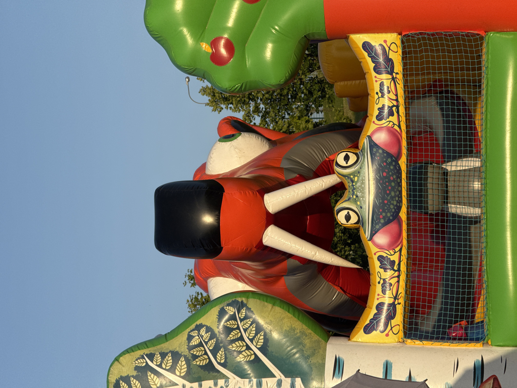 An outdoor, slightly low-angle, medium shot shows an inflatable bounce house with a sky background. The bounce house is red and gray with white tusks and a black head. It appears to be a dragon or some other mythical creature. To the left of the dragon is an inflatable tree with green leaves and white bark. To the right is a green inflatable apple tree with red apples. In the foreground is a frog's head with large eyes, red cheeks, and a colorful floral design. A green net separates the frog from the interior of the bounce house, which is red and yellow. Trees and a lamppost are visible in the background.