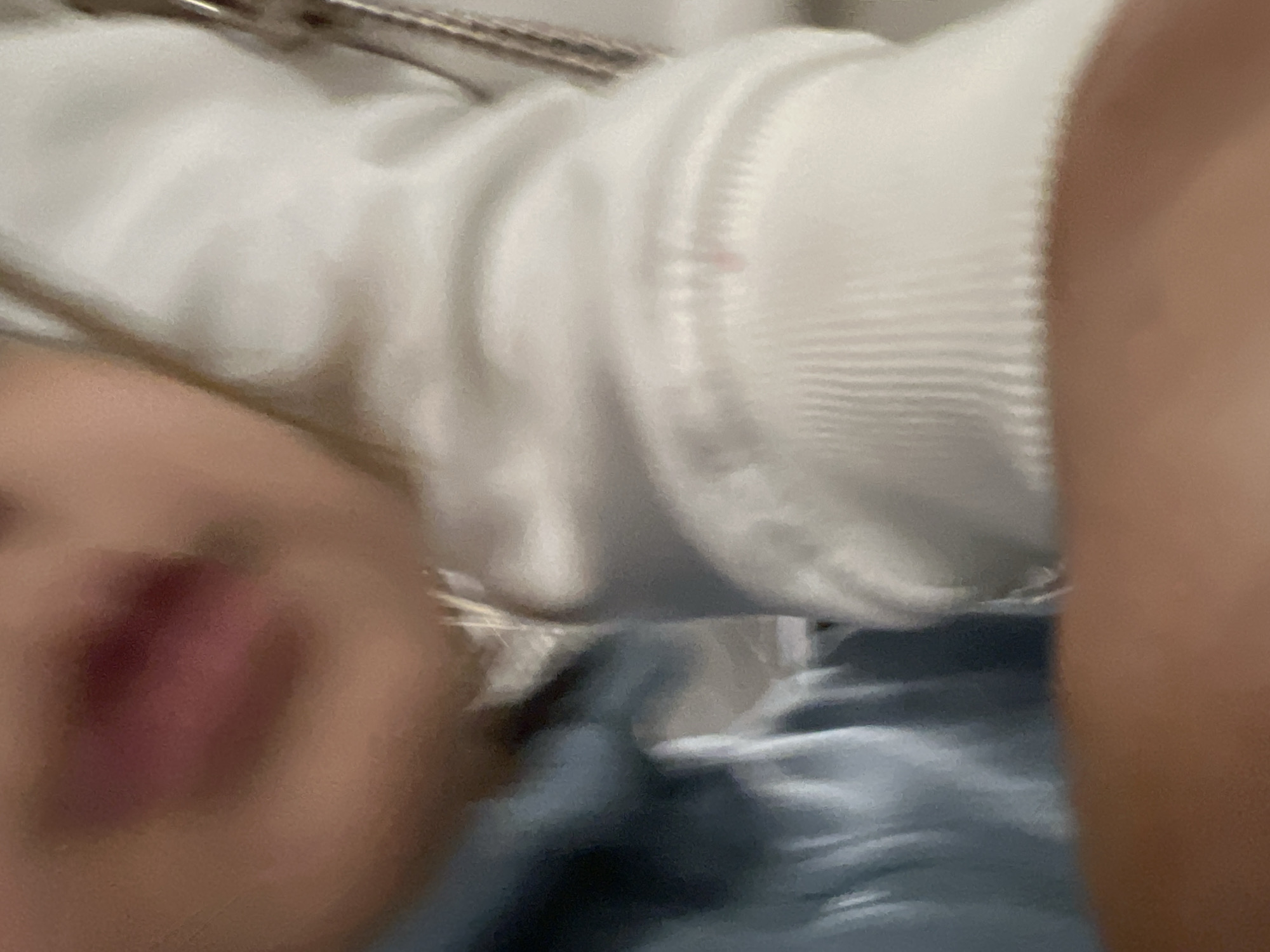 The image captures a heavily blurred, close-up perspective of an unidentified individual in Budva, Montenegro. The central focus is on the lower facial area, displaying lips with a dark pink or reddish hue. To the immediate left of the face, a portion of a light blue garment is visible. To the right, a white, ribbed sleeve, consistent with a sweater or hoodie, covers an arm. A small, faint pink mark is observed on the white sleeve above the cuff. The background is indistinct and out of focus, suggesting an interior setting. A metallic or reflective linear object is partially visible on the far right edge of the frame. No specific actions or interactions are discernible; the individual appears in a static pose.