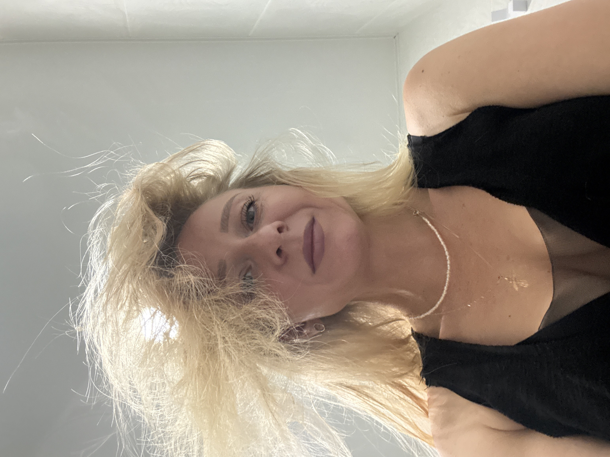 An outdoor, low-angle, waist-up medium shot captures a woman with blonde hair styled in a way that it appears to be blowing in the wind. She is wearing a black sleeveless top with a sheer mesh neckline, a pearl necklace, and a gold cross necklace. The background is a plain, light gray wall.
