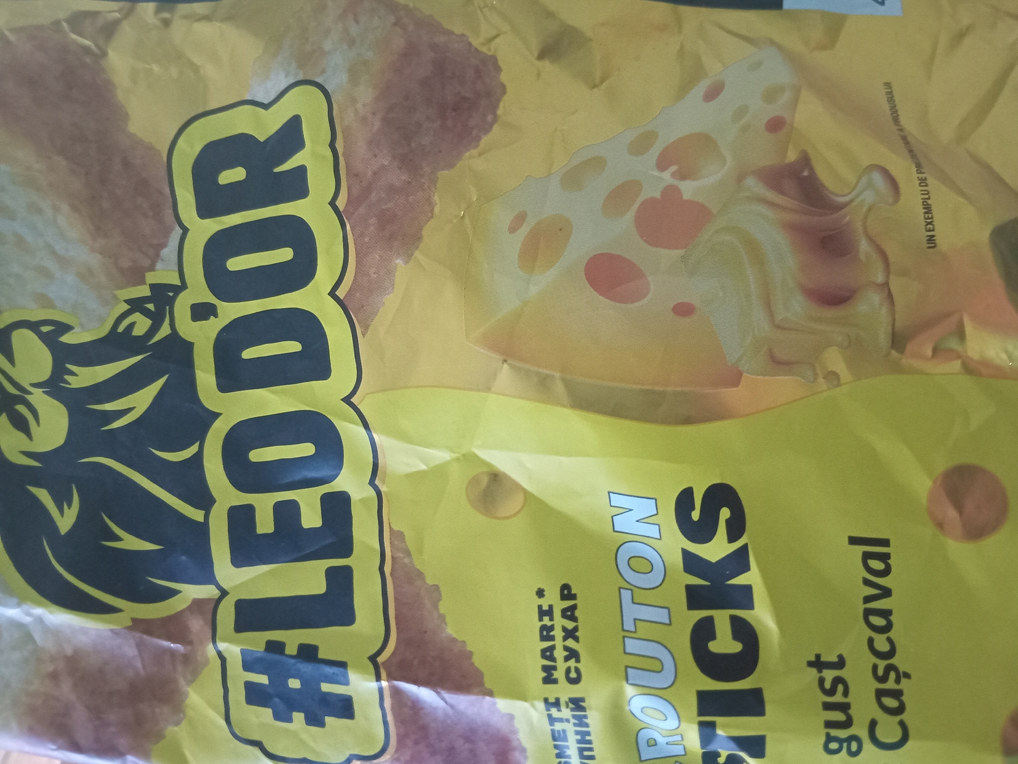 The image displays a close-up view of a crumpled yellow food packaging, likely for snacks. Dominating the upper portion of the package is a large graphic of a lion's head in black with yellow outlines. Below the lion, the hashtag "#LEODOR" is prominently displayed in large, black, stylized letters with a yellow outline. To the left of this text, some smaller text is visible, including what appears to be "SMETI MARI *" and "УПНИЙ СУХАР" in Cyrillic script. Further down, the words "ROUTON STICKS" are printed in large, silver-grey, sans-serif font. Below this, the words "gust" and "Casacaval" are written in a smaller, dark font, with "Casacaval" featuring a cedilla under the 'C'. The lower right section of the packaging shows an illustration of a slice of Swiss cheese with melting cheese. In the very bottom right corner, a line of very small text reads "UN EXEMPLU DE PREZENTARE A PRODUSULUI". The lighting is uneven, suggesting the package is indoors under artificial light, and the crumpled nature of the material indicates it's a flexible bag. There are no people or discernible environmental details beyond the product packaging itself. The location of Cotiujenii Mari, Moldova, is not depicted or suggested in the image content.