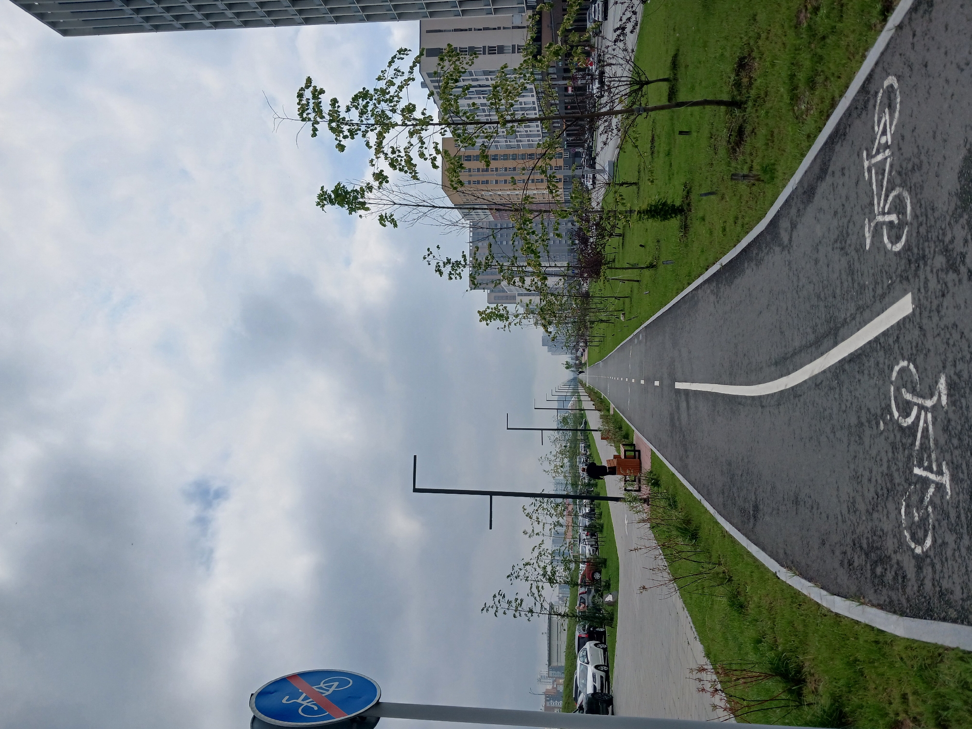 The image depicts a paved bicycle path in a modern urban environment. The path is marked with white lines, indicating separate lanes, and features bicycle symbols painted on its surface. Trees are planted along the sides of the path, separating it from a grassy area and a sidewalk. Beyond the grassy area, a parking lot filled with cars is visible, and further in the background, several multi-story residential buildings are present. A tall, modern building with a grid-like facade is also visible on the right side of the frame. The sky is overcast with clouds. A traffic sign indicating the end of a no-cycling zone is positioned on the left side of the path. A lone person is walking on the path in the middle distance. The scene appears to be in the city of Yekaterinburg, Russia, specifically in a developed residential area with infrastructure for cycling.