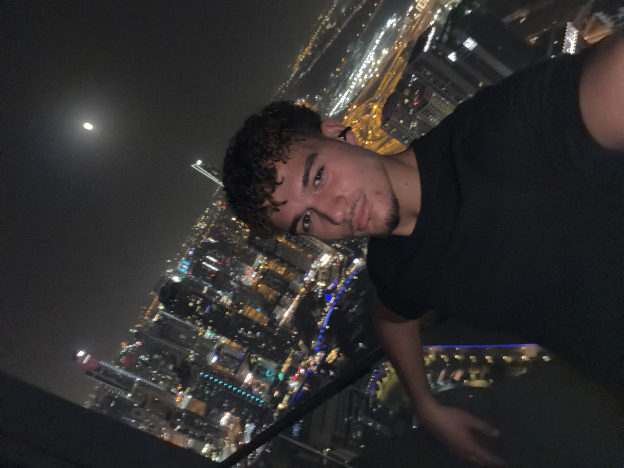 A person is photographed at night against the backdrop of the Dubai skyline. The person, positioned to the right of the frame, has curly hair and is wearing a dark-colored shirt. In the background, numerous buildings and streetlights create a dense, brightly lit cityscape. A diffuse light source, possibly the moon, is visible in the upper left portion of the sky. A dark structure, likely a ledge or railing, is visible in the lower portion of the frame, suggesting the photograph was taken from an elevated position. The scene is illuminated by the city lights, with the person’s face and upper body receiving sufficient lighting to be clearly visible. The general ambience is one of nighttime urban activity.