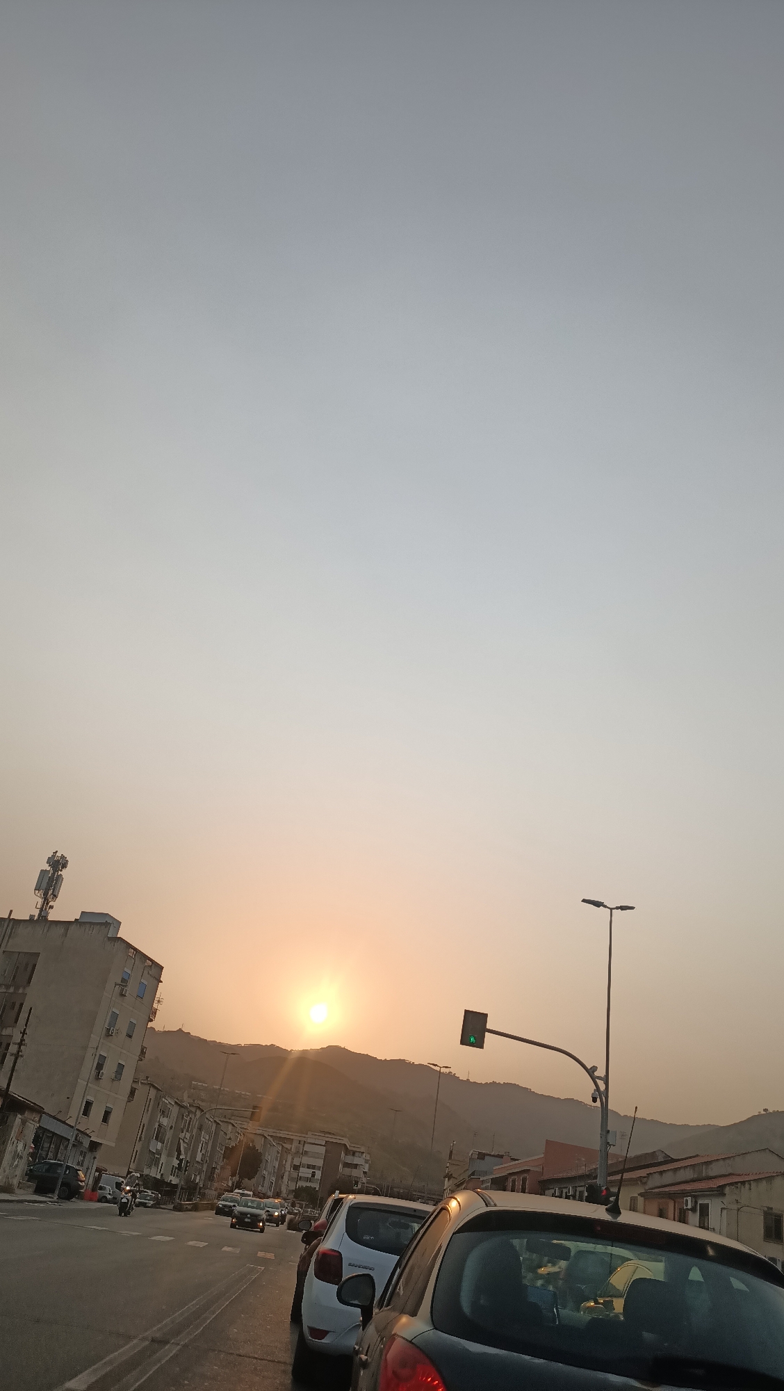 The image depicts a street scene in Messina, Italy, during a sunset. The sun, low on the horizon, casts a warm glow over a mountainous backdrop. In the foreground, several cars are visible on a road, suggesting traffic. Buildings line the street, some appearing to be apartment complexes. A traffic light with a green signal is present, indicating ongoing traffic flow. A cell tower stands atop one of the buildings on the left. The overall atmosphere is that of a typical urban evening, with the natural beauty of the sunset enhancing the cityscape.
