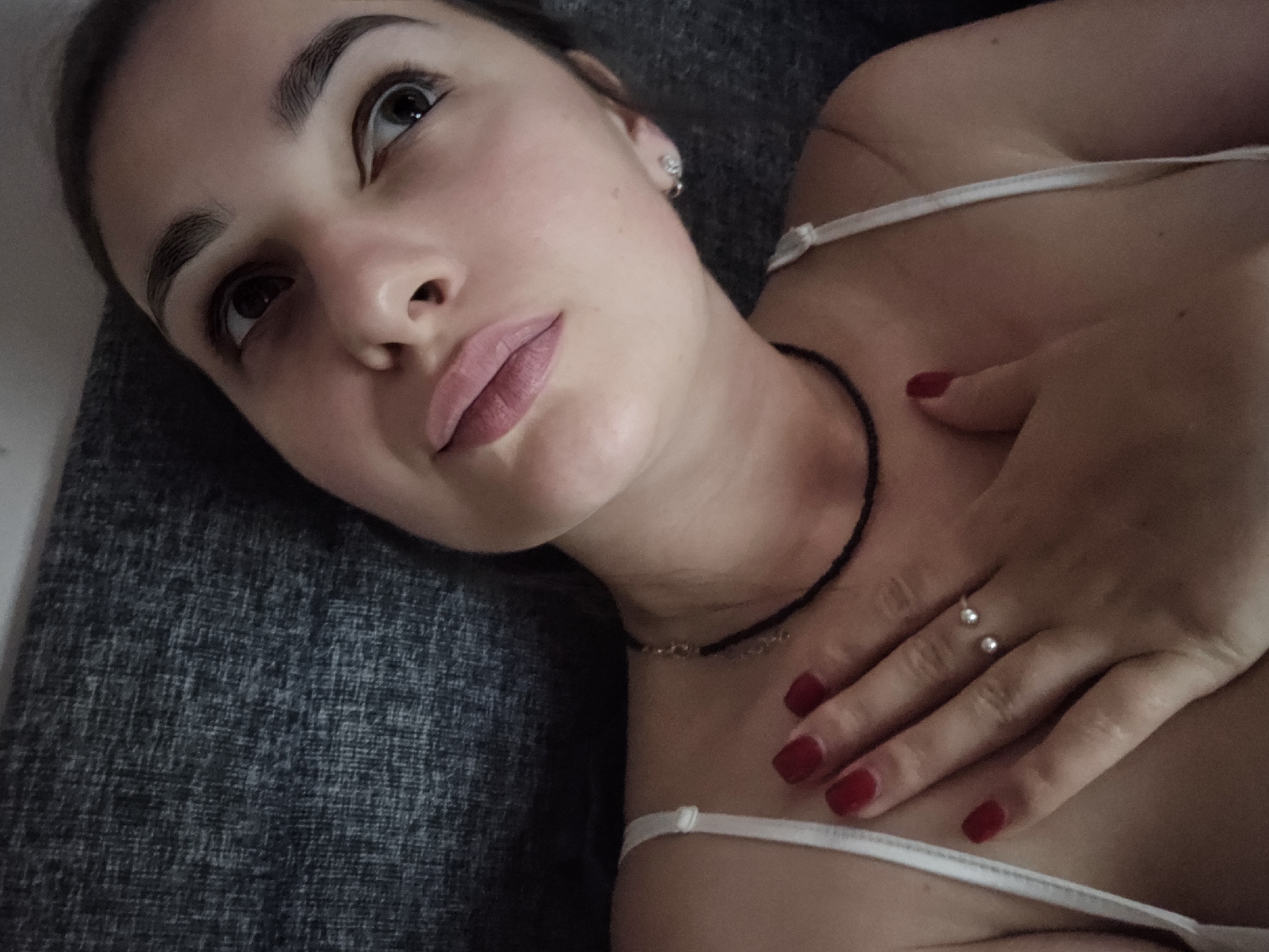 A young woman lies on a dark gray textured surface, her head tilted upwards and to the right. She has fair skin, dark eyebrows, and is wearing light pink lipstick. Her eyes are open and looking upwards and to the right. A thin black choker necklace is visible around her neck. She is wearing a white strap, likely from a camisole or bra. Her left hand, with bright red nail polish, rests on her chest, with a delicate silver ring with two small pearls on her ring finger. A small, diamond-like stud earring is visible on her right ear. The lighting is dim, casting soft shadows. There is no information about the city of Petropavlivska Borshchahivka, Ukraine, in the image.