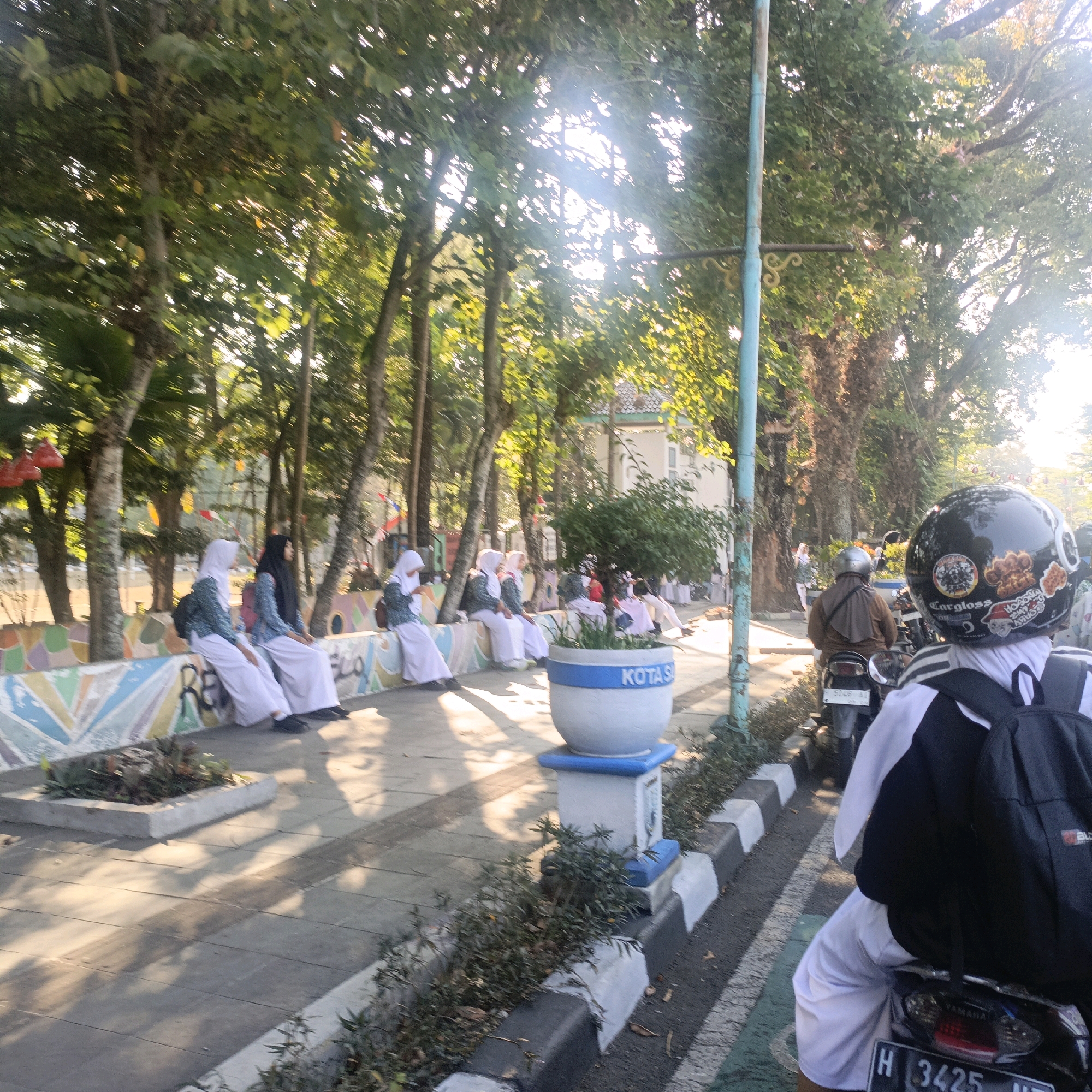 In Salatiga, Indonesia, on a tree-lined street, several young women in white hijabs and uniforms are seated on a decorative concrete wall. Sunlight filters through the dense foliage of mature trees, casting dappled shadows on the pavement. A large white planter with "KOTA SA" visible on its side stands nearby. In the foreground, a person wearing a black helmet decorated with stickers and a black and white backpack sits on a motorcycle, facing away from the camera. The motorcycle's license plate reads "H 3425 NT". The scene appears to be a public walkway or roadside area.