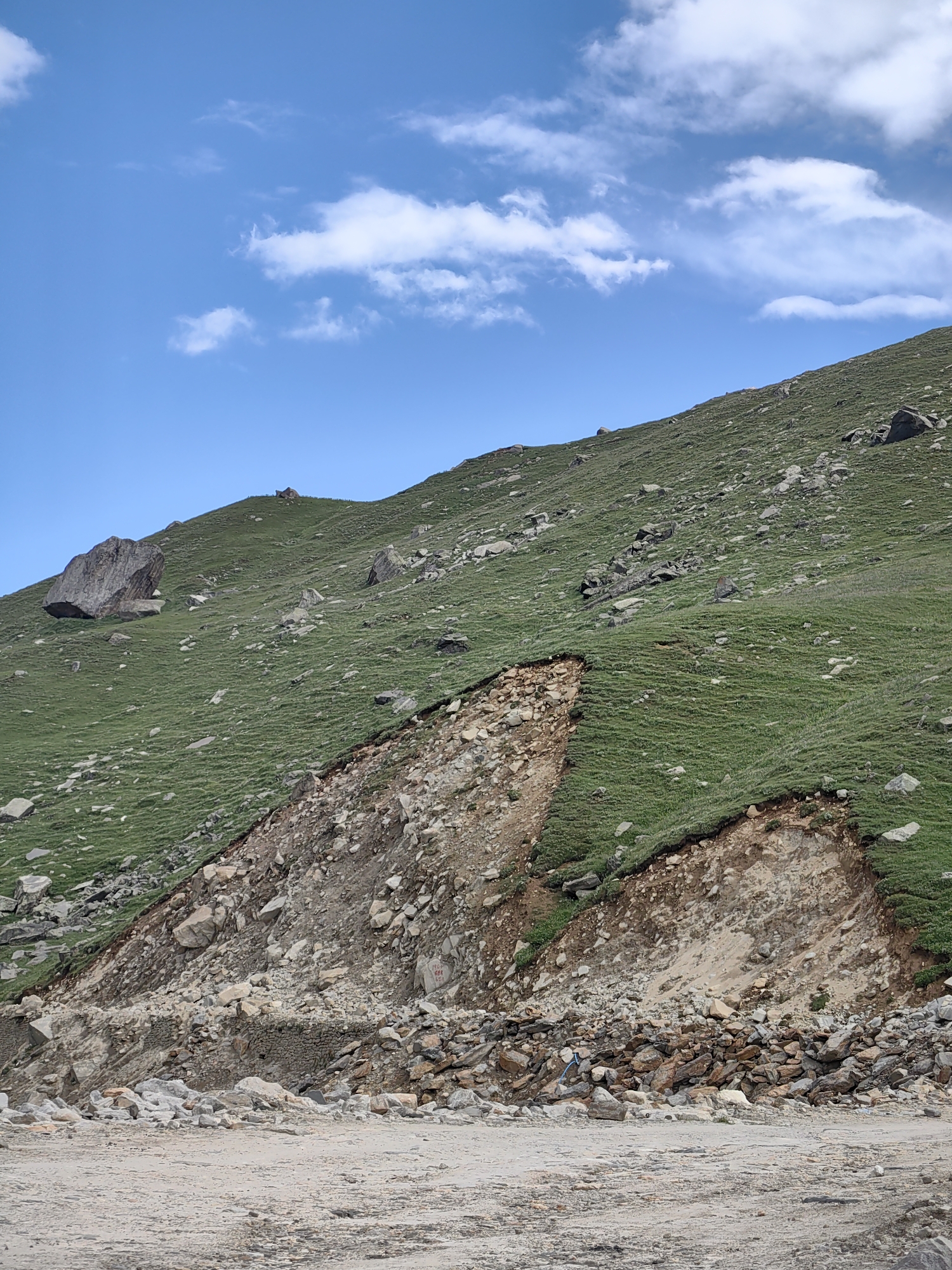 A wide-angle, low-angle shot captures a steep, grassy hillside under a bright blue sky dotted with white clouds. The hillside is covered in short, vibrant green grass and scattered with numerous grey and brown rocks of varying sizes. Towards the bottom of the frame, the hillside gives way to a steep, rocky embankment with exposed soil and a landslide scar. Several large rocks are visible in this area, some appearing to be recent fall. The ground in the foreground is a mix of dirt and small stones. The image appears to be taken in a mountainous region, likely in the vicinity of Bashisht, India, given the natural landscape. There are no people or animals visible in the scene.