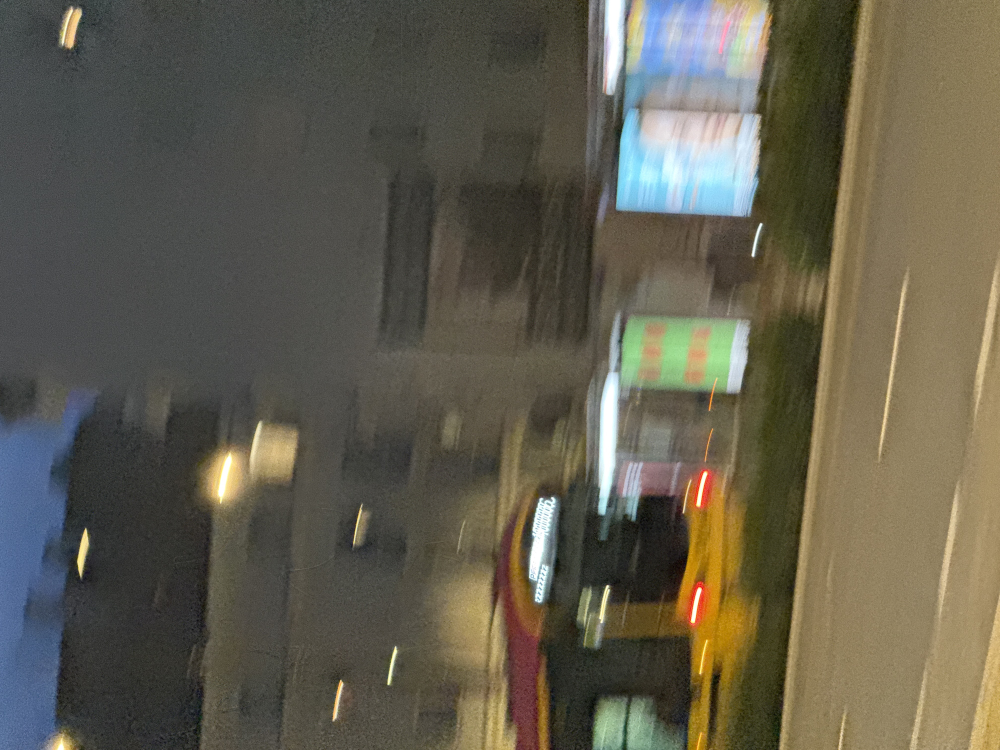 The image depicts a blurred nighttime scene on a city street. In the foreground, a road with white lane markings is visible. Behind the road, there is a strip of green foliage. Further back, a moving yellow vehicle with red taillights is partially visible. Above and behind the vehicle, a building with illuminated windows is present. In the middle ground, there are several large, blurred advertisements, one with a green background and another with a blue background. The sky above is a dark, twilight blue. The overall impression is one of motion and the ambiance of a city at night, likely captured from a moving vehicle.