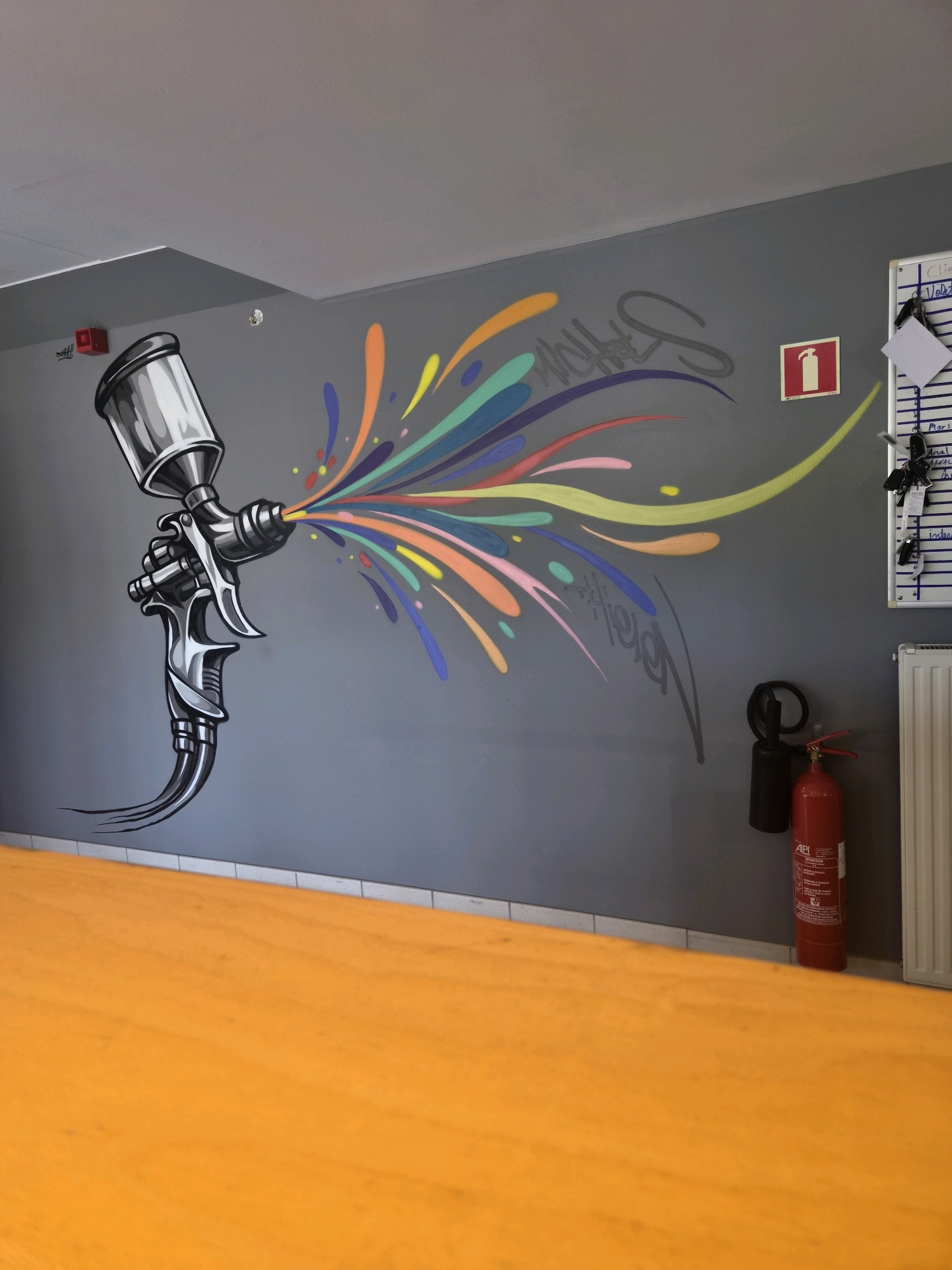 In Sanem, Luxembourg, a vibrant mural adorns a gray wall, depicting a chrome spray gun in the process of spraying a cascade of colorful paint splatters and arcs. The artwork is detailed, with shading that gives the spray gun a metallic sheen and the paint a dynamic, energetic quality. To the left of the mural, a red fire alarm is mounted on the wall. To the right, a whiteboard with handwritten notes and keys hanging from it is visible, along with a red fire extinguisher and a black hose reel attached to the wall. A white radiator is situated below the whiteboard. The lower portion of the image is dominated by a bright orange surface, likely a table or counter, which contrasts sharply with the gray wall and the mural. The lighting suggests an indoor environment, possibly a workshop or studio.