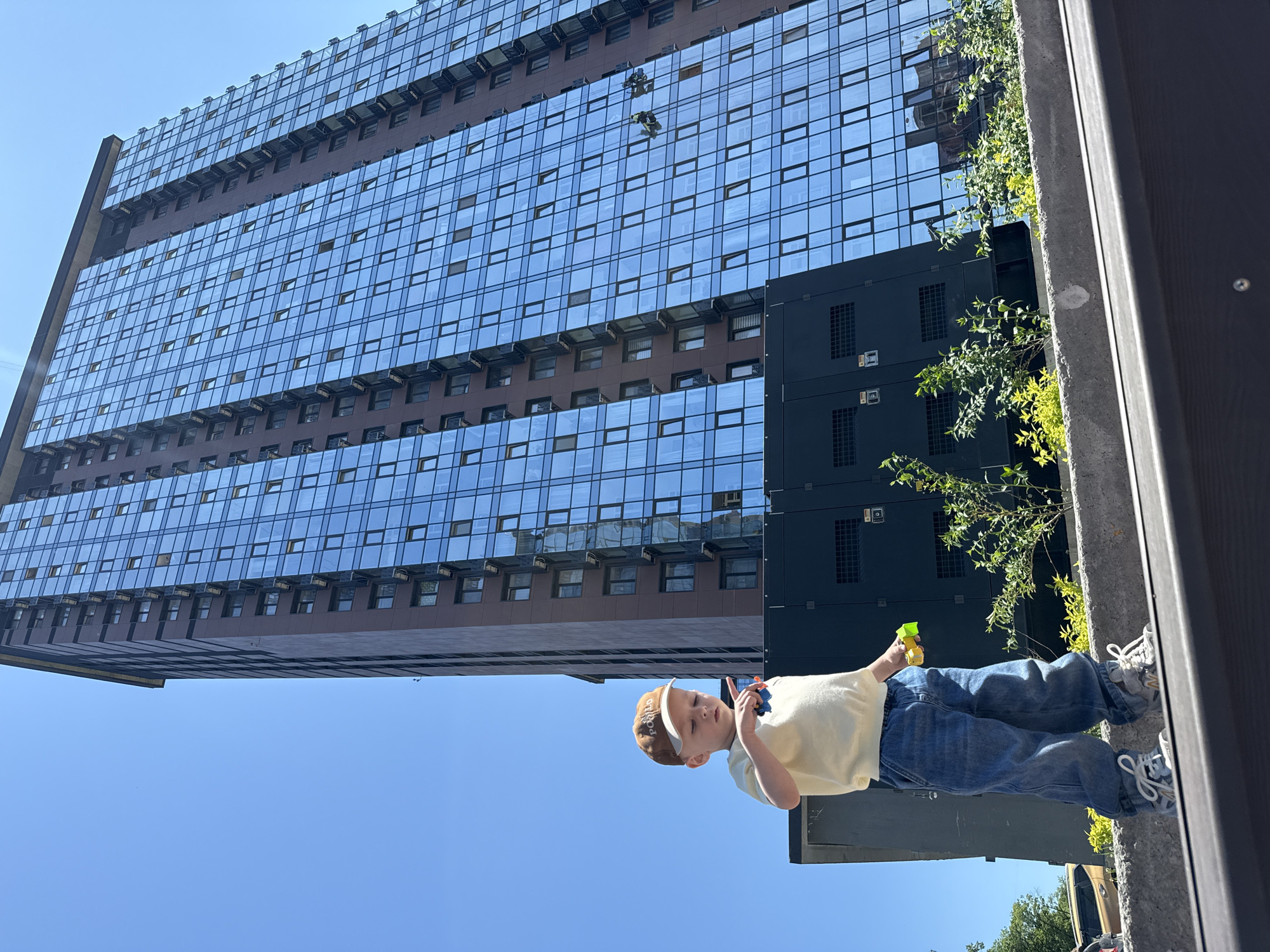 A child stands in front of a tall building, likely in Vladivostok, Russia, given the architectural style of the building and the Cyrillic lettering on the boy's cap. The building is a modern high-rise with a facade of mirrored glass windows set in a brown grid. Large black boxes are fixed to the building's side. Green foliage grows from a ledge near the black boxes. The child is wearing jeans, a light-yellow t-shirt, a baseball cap, and sneakers. He is holding a small toy or object in his hand. The sky is clear and blue. The photograph has been rotated 90 degrees counterclockwise.