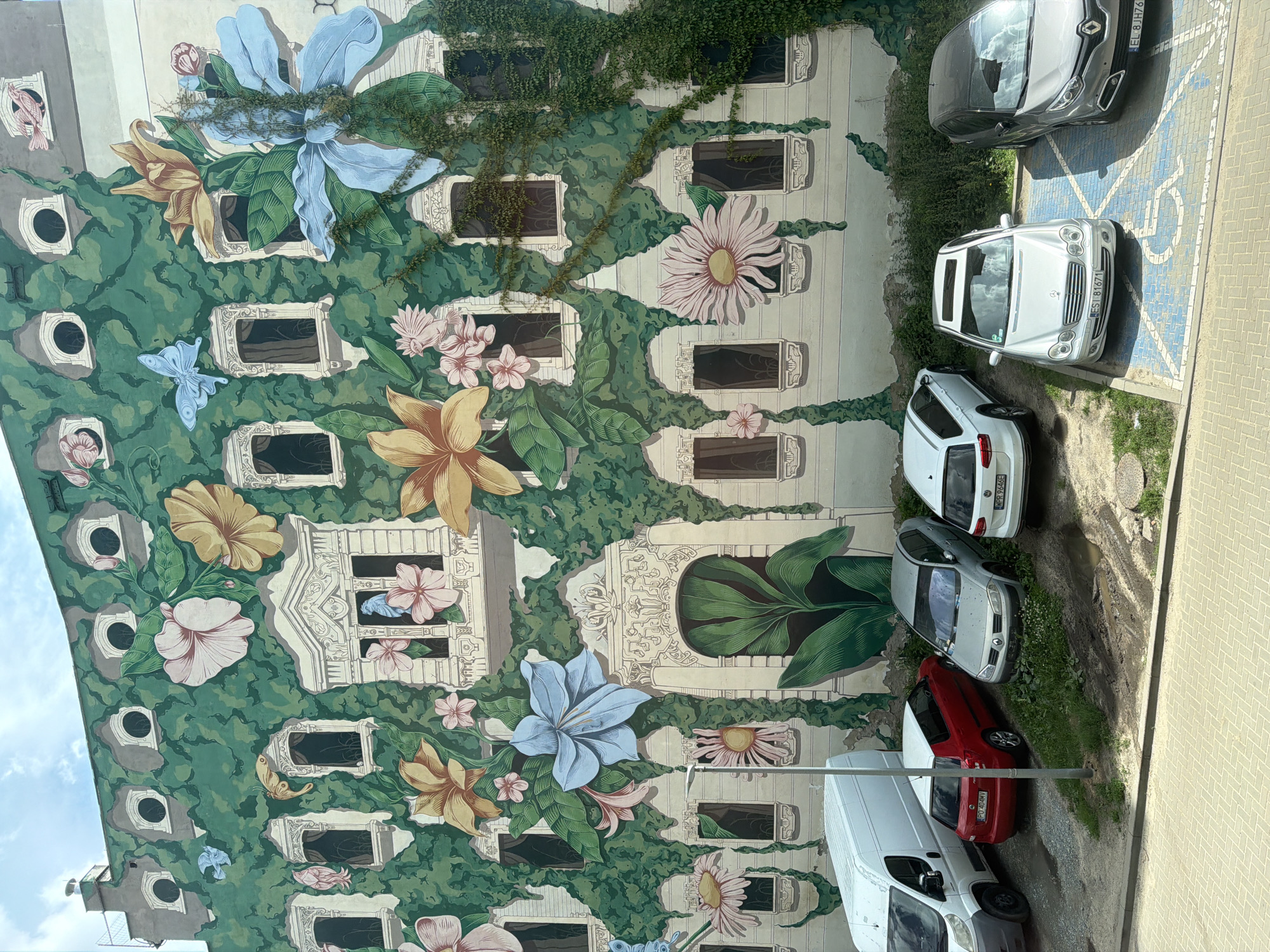 The image is a photograph taken from a high angle, looking down at a building with a large mural on its exterior. The mural depicts a whimsical scene with oversized flowers, vines, and windows. Various stylized flowers in shades of blue, pink, yellow, and white are scattered across the green, vine-covered facade. Several windows are integrated into the mural, appearing as framed openings within the painted design. A few small butterflies are also depicted.
In the lower right portion of the image, there is a parking area with several cars. These include a silver Renault, a white Mercedes-Benz, a white SUV, a red car, and a white van. The cars are parked on a paved surface adjacent to a patch of grass and dirt.
The time of day appears to be daytime, with sunlight casting shadows. The sky is visible in the upper left corner and appears cloudy. The overall impression is a blend of urban art and everyday life, with the vibrant mural contrasting with the parked vehicles and the utilitarian building structure. There are no discernible people or specific events taking place. The location is unknown, but the presence of cars and a building suggests an urban or suburban setting. Some of the visible license plates are partially obscured or illegible, but one appears to read "EL 8JH761" and another "ES1 B1671".