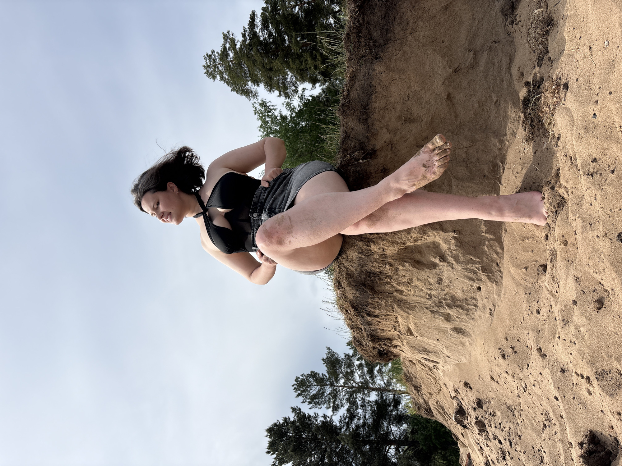 A woman sits on a sandy bank in what appears to be an outdoor setting. The location is specified as Chaikovsky, Russia.
The woman is wearing a black swimsuit top and grey shorts. Her legs are crossed, and her feet are bare and covered in sand. She is looking slightly downward and appears to be smiling.
The setting includes a sandy bank or cliff, with visible soil layers and some sparse vegetation. Trees are seen in the background against a light sky.