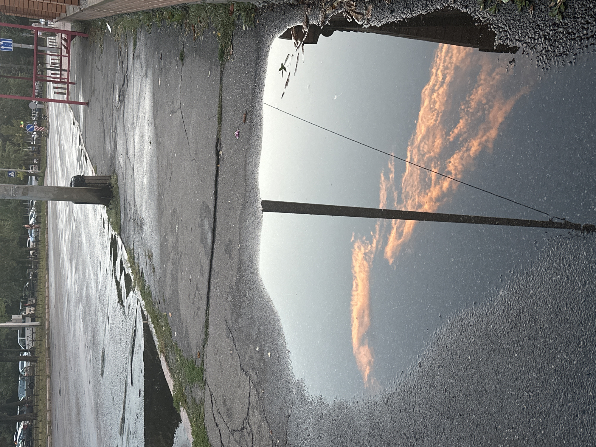 The image depicts a puddle on a city sidewalk in Kyiv, Ukraine, reflecting the sky and surrounding environment. The puddle is located on a cracked asphalt sidewalk adjacent to a street with parked cars and trees. A tall gray pole stands vertically in the puddle, bisecting the reflection. The reflection shows a sky with orange-tinged clouds, suggesting it might be sunset or sunrise. A red metal structure, possibly part of a bus stop shelter, is visible on the sidewalk further down. The ground around the puddle is composed of asphalt and gravel. The overall scene suggests a wet, post-rain or damp urban environment.