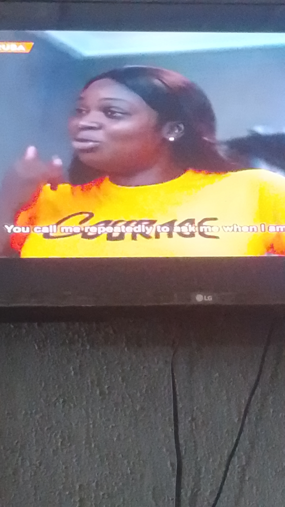A woman with dark hair and a bright yellow shirt that reads "COURAGE" is speaking, her hand raised. The text on the shirt is in black lettering with a fiery red and orange outline. The background is a blurred, greyish-blue, and a small orange logo with the word "LUBA" is visible in the upper left corner. A faint "LG" logo is visible on the bottom edge of the frame, suggesting the image was taken from a television screen. The woman appears to be in an indoor setting, possibly a studio or a living room, as indicated by the television screen. The overall scene is a close-up shot of the woman and her shirt.