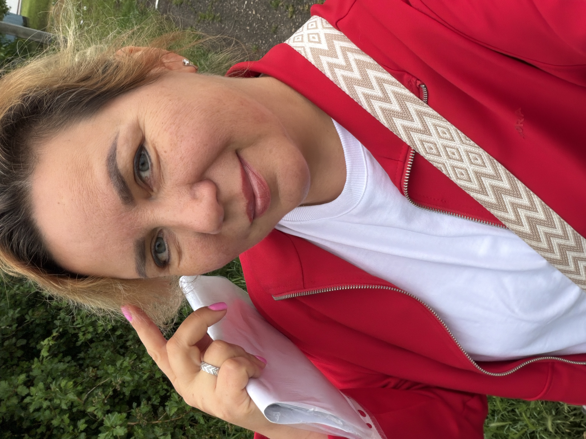 A woman is holding a piece of paper. She is wearing a red jacket and a white shirt. The woman's fingernails are painted pink. The woman is wearing a ring on her left ring finger. The environment appears to be outdoors, with some grass and greenery visible in the background. It is not possible to determine a location in the city of Leningradskaya, Russia, with the given information.