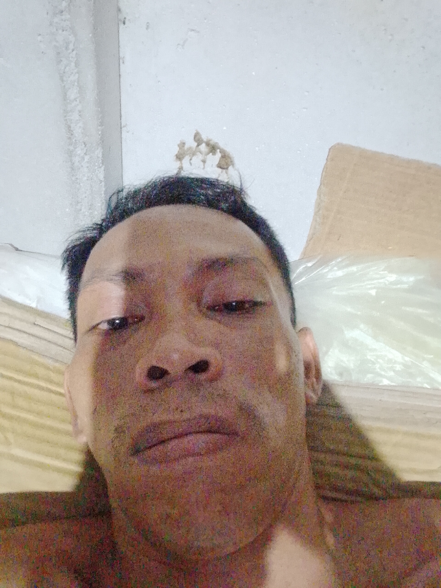 A close-up selfie of a middle-aged Asian man is presented. He has short, dark hair and a neutral expression. He is lying down. Behind him is a white wall, cardboard boxes and a translucent plastic bag. The location is Bukit Ketok, Indonesia.