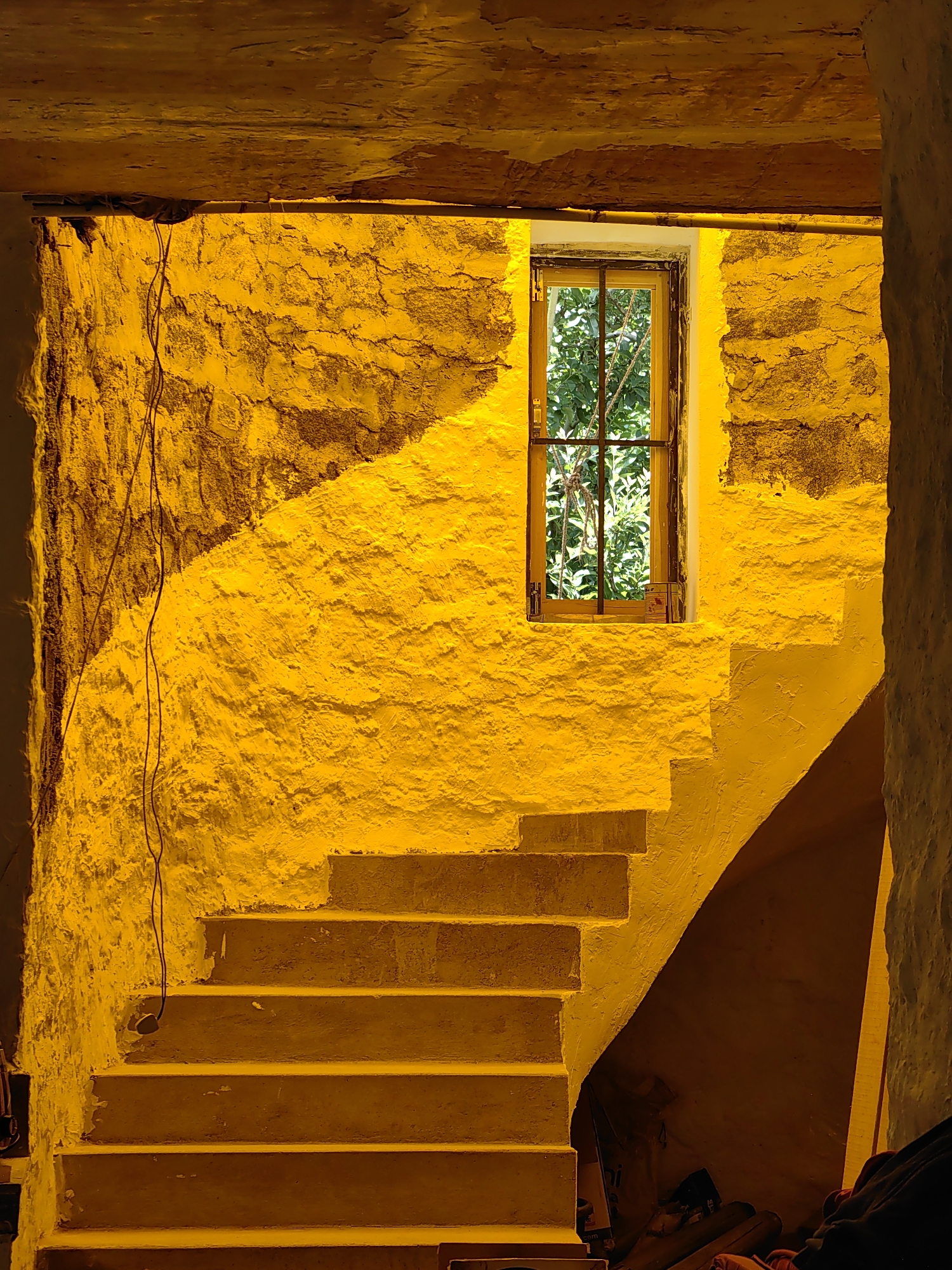 The image depicts an interior view of a stone building with a staircase in Vashisht, India, illuminated by a warm, yellow light, likely from an artificial source. The rough, textured stone walls are painted white and are partially illuminated by a strong diagonal beam of light coming from a window. The window, with a metal grate, looks out onto lush green foliage. A set of concrete stairs ascends from the lower left of the frame, curving to the right. The lower part of the stairs is in shadow, while the upper steps are brightly lit. Wires hang down the left wall. In the lower right corner, the space beneath the stairs is dark and contains indistinct objects that appear to be stored items. The overall impression is one of rustic architecture and dramatic lighting within an aged structure.
