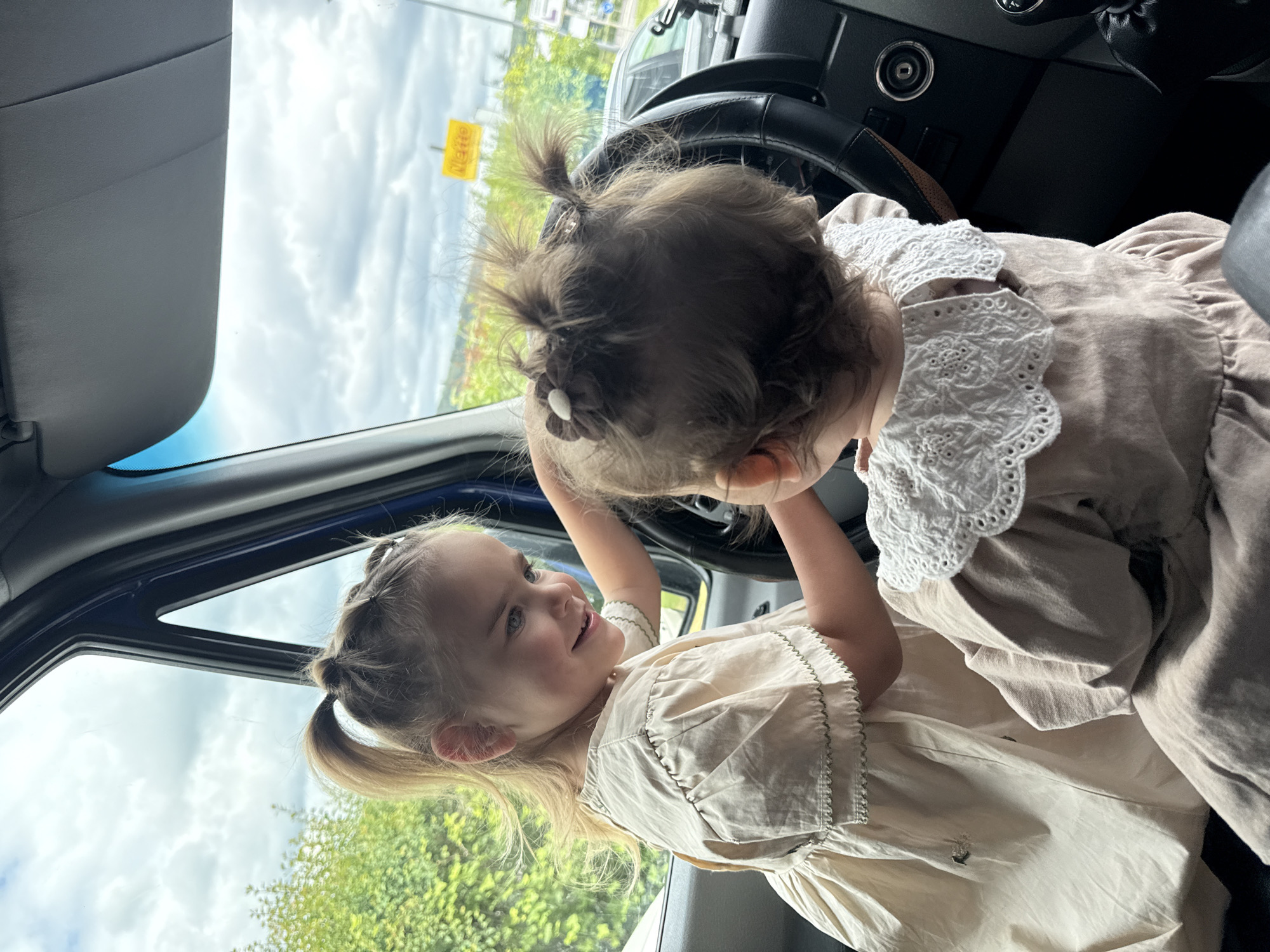 Two young girls are seated inside a vehicle, with one girl positioned in front of the steering wheel, appearing to be driving. The girl in the driver's seat is seen from the back, with her dark hair styled in multiple small ponytails and a decorative bow. She is wearing a light-colored garment with a white lace collar. The second girl, facing the driver's seat, has blonde hair tied in a ponytail and is wearing a light-colored dress with green trim. She is smiling and reaching towards the steering wheel. Outside the vehicle, a cloudy sky and green trees are visible, suggesting an outdoor setting. A yellow sign with red text is partially visible in the background. The scene is captured from a perspective inside the vehicle, looking towards the front.