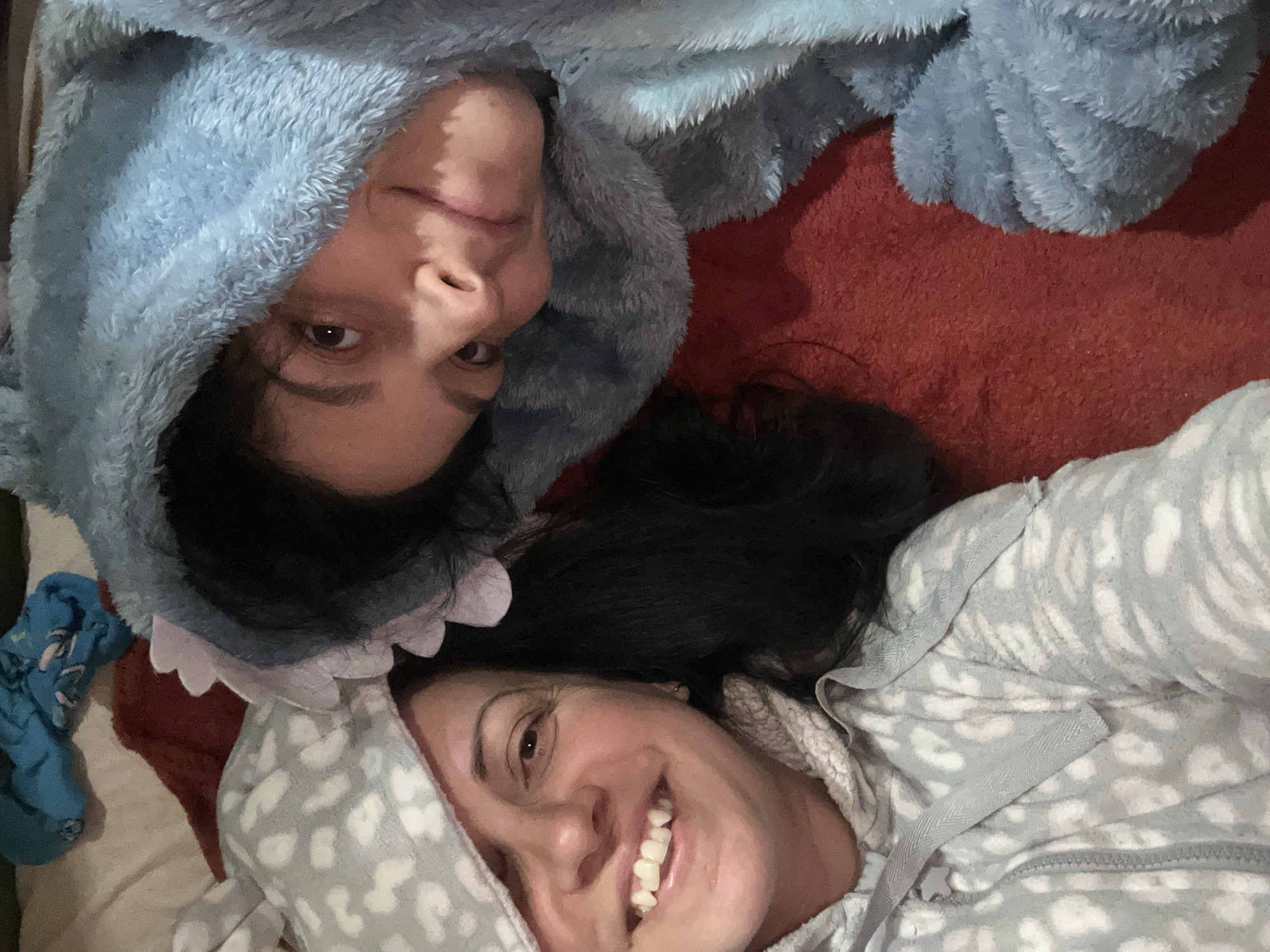 Two women are lying down side-by-side. The woman on the left is wearing a blue, fuzzy hooded shark onesie with white teeth along the hood's edge. The woman on the right is wearing a gray and white hooded pajama top with a leopard print. Both women are smiling. The background consists of a red blanket, and a blue sock can be seen in the bottom left corner of the image. The location is unknown; there is no indication that the photo was taken in Sofia, Bulgaria.