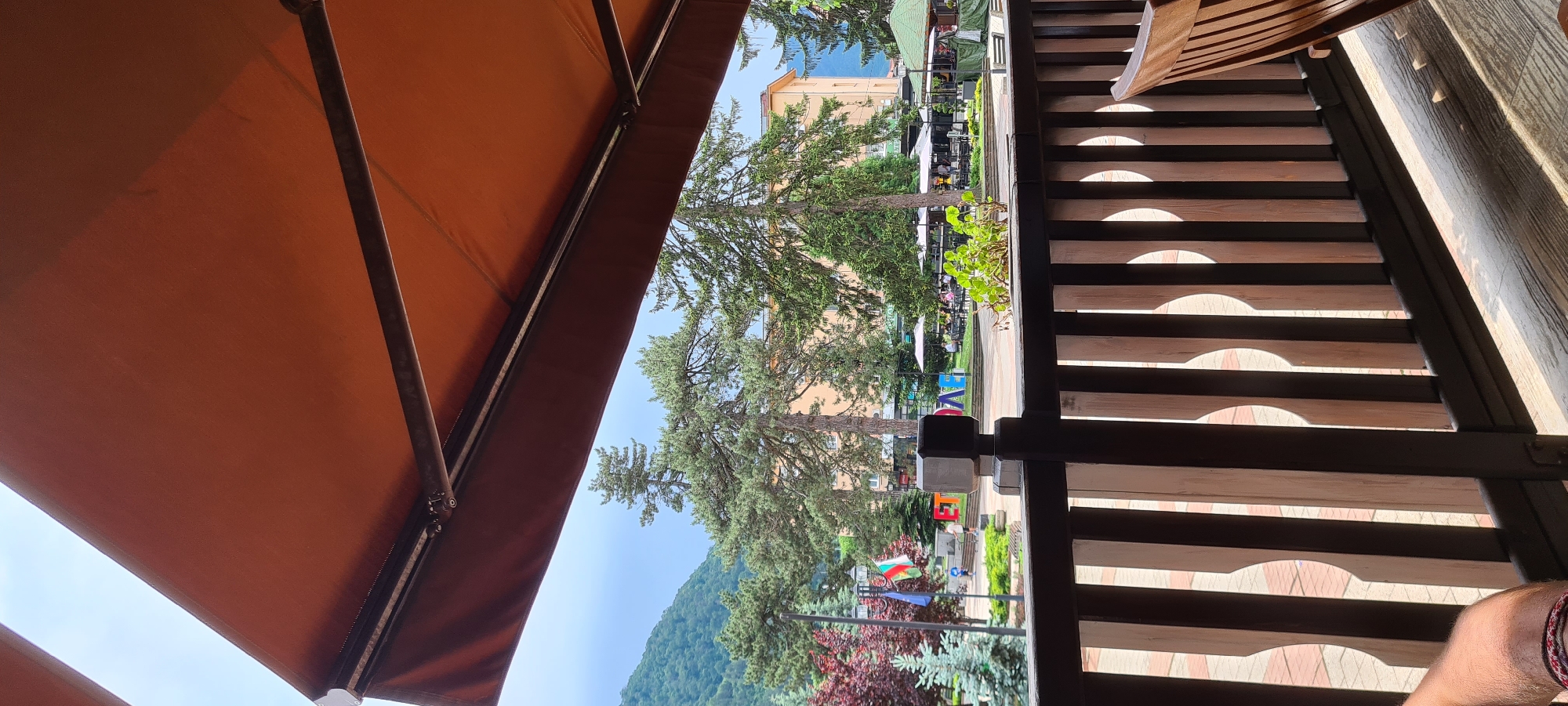 The image captures an outdoor scene in Etropole, Bulgaria, viewed from under a rust-orange retractable awning, likely from a cafe or restaurant patio. In the immediate foreground, the underside of the awning dominates the upper portion of the frame, showing its mechanical framework and fabric. A dark brown wooden balustrade runs horizontally across the lower mid-ground, featuring vertical slats that cast shadows on the floorboards below. Part of a person's left forearm, adorned with a woven bracelet, is visible in the bottom-left corner, resting on a surface. A portion of a light brown wooden chair back is present in the bottom-right.
Beyond the balustrade, a paved town square is visible, populated by several individuals. To the left, a light pole displays a Bulgarian flag and a plain blue flag. Further into the square, large, multicolored block letters spell out "ЕТРОПОЛЕ" (Etropole). Several people are present, with some individuals walking in the square and others seated at tables under white umbrellas, indicating an outdoor cafe area to the right. Greenery, including tall pine trees and smaller red and green bushes, is distributed throughout the square. Buildings, predominantly light yellow-orange, are visible behind the square. In the distant background, forested mountains rise under a light blue sky with scattered clouds. The overall lighting suggests daytime conditions.