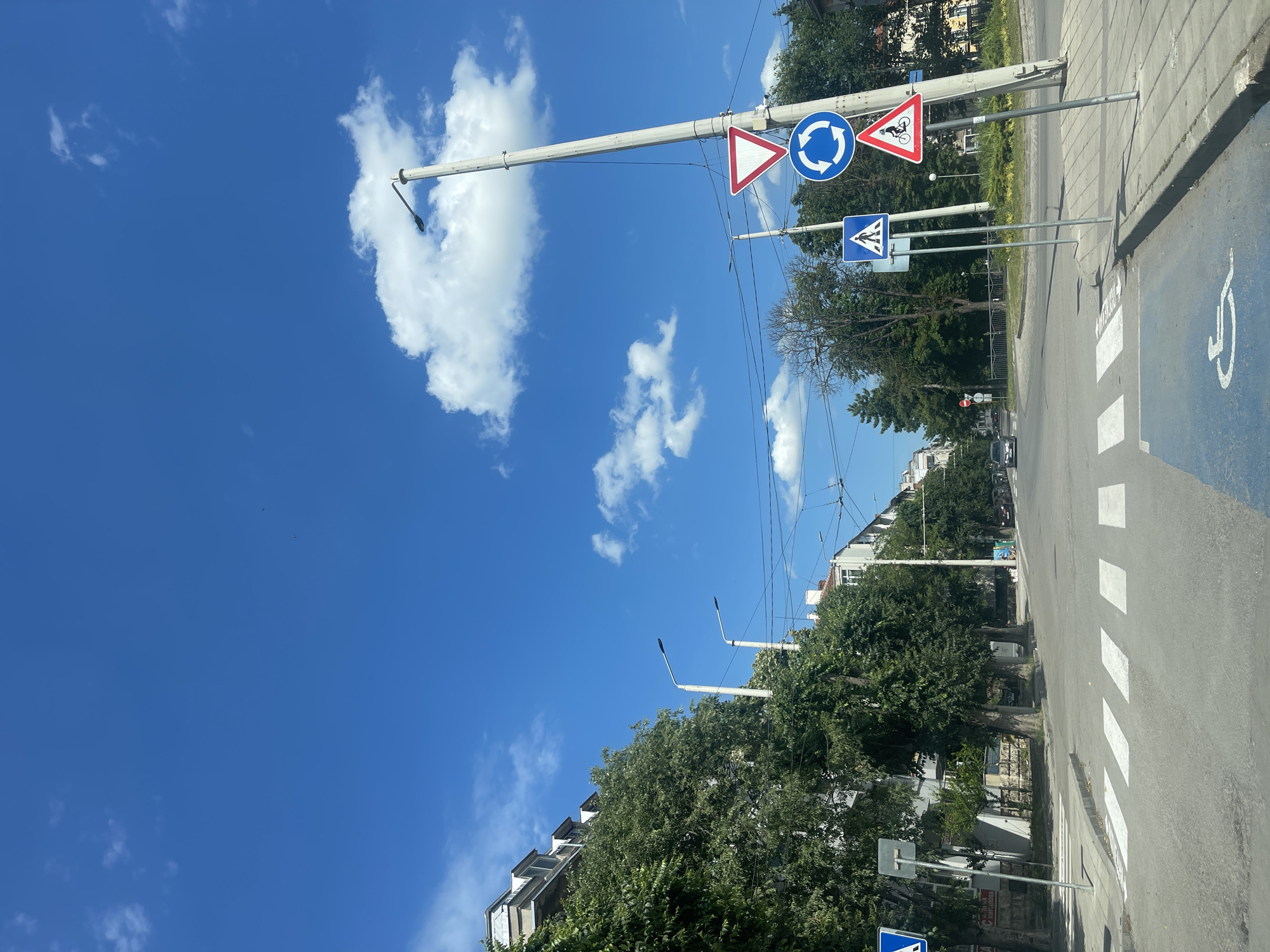 The image depicts a street scene in Ruse, Bulgaria, likely near an intersection. The perspective is from a vehicle, looking forward towards the intersection and along a tree-lined street.
**Environment:**
* **Sky:** A bright blue sky with scattered white cumulus clouds.
* **Roadway:** Paved asphalt road with clear white pedestrian crossing markings (zebra crossing). A blue painted area with a white wheelchair symbol is visible on the road surface, indicating a designated disabled access point.
* **Sidewalk:** Concrete sidewalk bordering the road.
* **Buildings:** Multi-story buildings with light-colored facades line the street on the left.
* **Vegetation:** Numerous mature trees with dense green foliage are planted along the sidewalk and in medians, providing shade. Some bushes are also visible.
* **Street Furniture/Infrastructure:**
* Several lampposts are present, one of which is a tall, concrete utility pole supporting multiple traffic signs and overhead electrical wires.
* Traffic signs are prominent:
* A triangular red-bordered "Give Way" sign.
* A circular blue sign with white arrows indicating a roundabout.
* A triangular red-bordered sign depicting a cyclist.
* A blue square sign with a white pedestrian symbol.
* Another blue sign with a white arrow on the left side of the road.
* Overhead electrical wires are strung across the street, likely for trams or public transport.
* **Vehicles:** A few cars are visible in the distance on the road, and parked cars line the right side of the street.
**Action/Subjects:**
* No people are clearly visible and identifiable in the scene.
* The scene is static, portraying a typical urban street environment during daylight.
**Location Specifics:**
The presence of specific traffic signs (roundabout, give way, cyclist warning) and the overall architectural style of the buildings are consistent with a European city. Without additional landmarks or text, pinpointing the exact location within Ruse is not possible from the image alone, but it suggests an area with residential and commercial buildings, as well as significant green spaces. The tram wires suggest a city with a tram system.