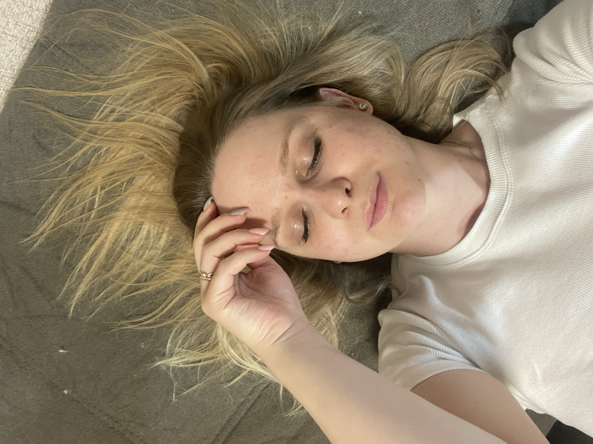 The image shows a close-up, overhead view of a young Caucasian woman lying on a textured, dark grey surface, possibly a sofa or bed. Her eyes are closed, and her right hand is resting on her forehead. She has blonde hair, which is spread out around her head, and is wearing a white, ribbed t-shirt and a delicate necklace. Her skin appears fair with some minor blemishes. There are a few small white specks visible on the grey surface near her hair. The overall impression is one of rest or relaxation. The image does not contain any information or visual cues that would allow for its location in the city of Chaikovsky, Russia.