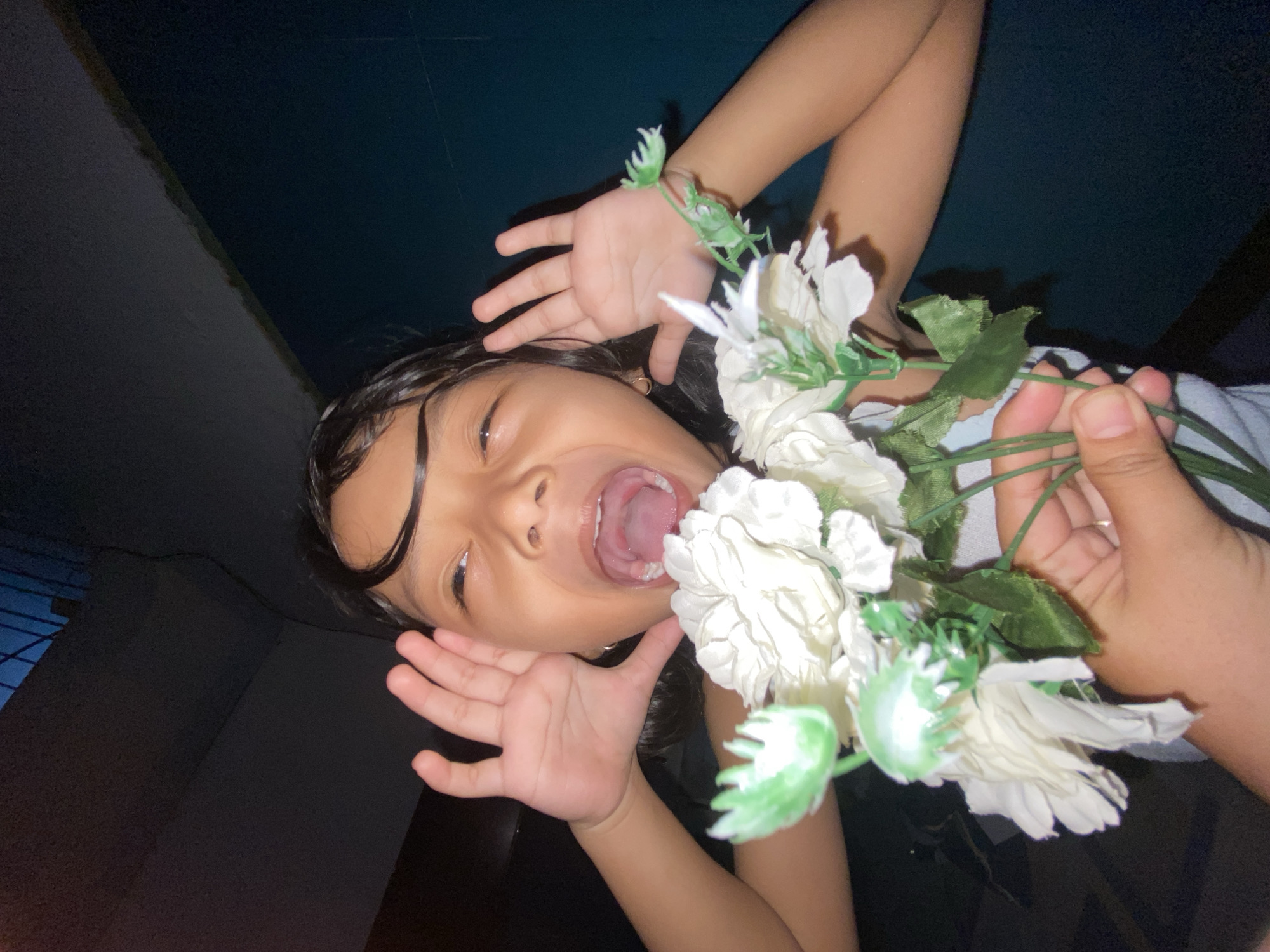 A young person, with dark hair and tan skin, is holding a bouquet of white flowers. They are positioned at an angle to the camera, facing upwards with their mouth open wide. Their hands are raised on either side of their face. The background is dark, possibly an indoor space. The location is Gelam, Indonesia.