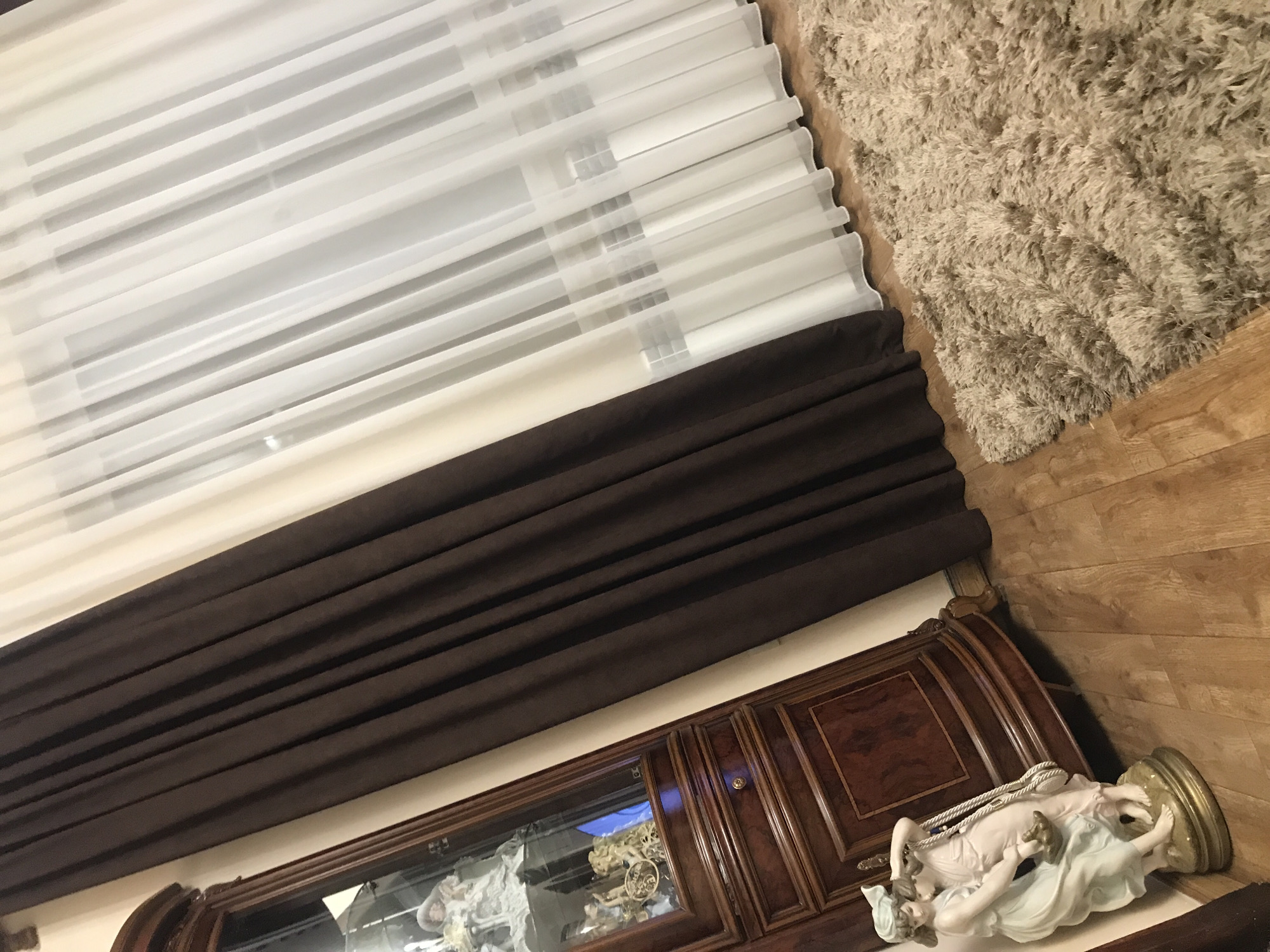 The image shows a room interior with a few notable elements. A set of white blinds is positioned at the top left. Beneath the blinds, there's a dark brown curtain or fabric covering a wall. Below the curtain, a wooden cabinet with a glass display area is visible. Inside the cabinet, some figurines or decorative items are present.
To the right, a beige rug covers a section of the floor. Next to the rug, the wooden floor is exposed. On the right side of the image, resting on the floor next to the cabinet, is a porcelain statue of a woman.
The location is not discernible from the image. The description does not suggest that the image is necessarily taken in Yerevan, Armenia, or anywhere else.