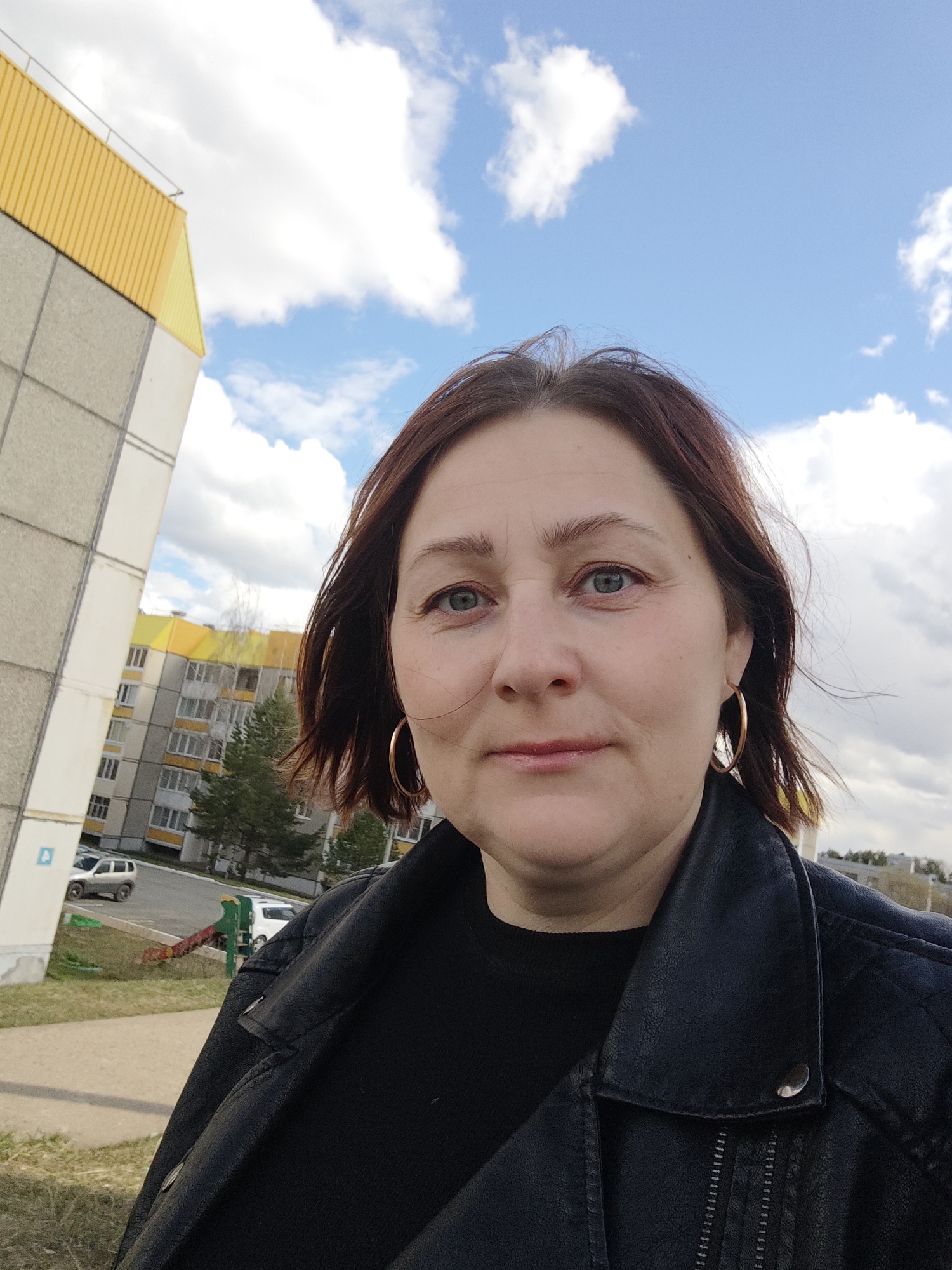 The image is a daytime, outdoor portrait of a woman looking directly at the camera. She appears middle-aged, with reddish-brown, shoulder-length hair, blue eyes, and a neutral to slightly smiling expression. She is wearing a black leather jacket over a black top and has gold-colored hoop earrings. Her hair is slightly windblown.
The background features a bright blue sky filled with scattered white, fluffy clouds. To the left, a large, multi-story building with a distinctive yellow upper section and grey and white paneling dominates the frame. A small blue rectangular sign with a white symbol (possibly a letter or number) is visible on its lower section. In the mid-ground, another multi-story residential building with yellow accents is visible behind trees, some of which are bare-branched, along with a prominent coniferous tree. A paved path or sidewalk, patches of green grass, and several parked cars (including a light-colored SUV and a white car) can be seen further in the background. The scene suggests an urban or suburban residential area, consistent with the Марковский, Russia location, on a clear and possibly breezy day. There is no specific activity taking place other than the woman posing for the photo.