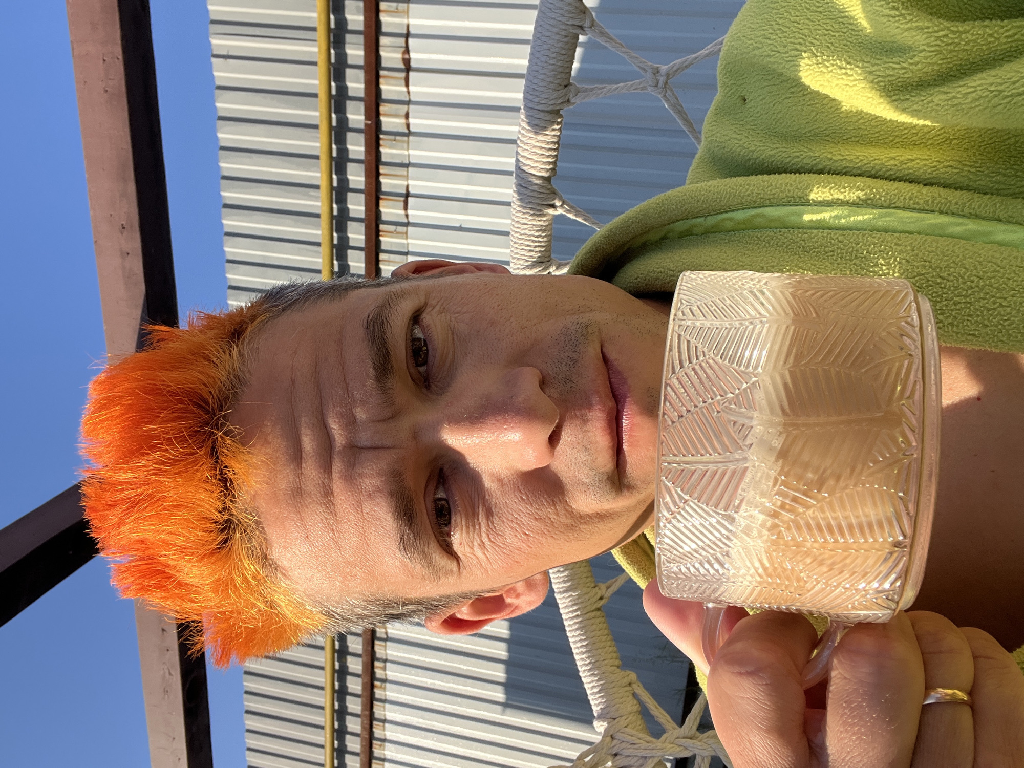 A man with brightly dyed orange hair and a gray robe holds a clear glass mug filled with a light-colored beverage, possibly coffee with milk, in front of a corrugated metal fence under a clear blue sky. The man appears to be outdoors, possibly on a balcony or patio, with a rope swing chair visible. The sun is casting shadows and highlights on his face and the mug. No specific location within Chaikovsky, Russia is identifiable.