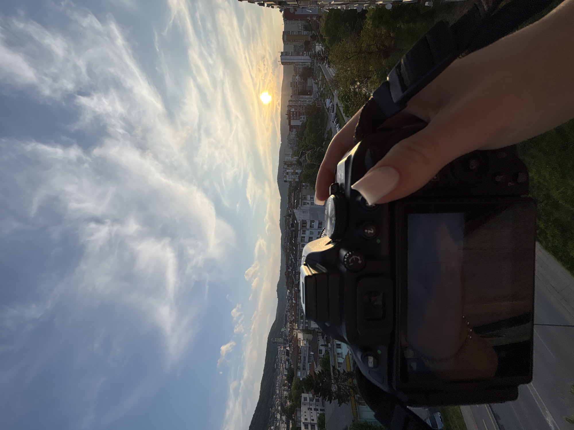 A medium shot, rotated clockwise 90 degrees, shows a woman's hand holding a black camera in the foreground against a cityscape at sunset in Unknown. The camera, occupying the right half of the frame, is a DSLR model with various buttons and dials visible. The woman's hand grips the camera body, with a visible nail on the index finger. The background reveals a cityscape with buildings of varying heights, a road, trees, and a setting sun. The sky is filled with scattered clouds and a golden hue from the setting sun.