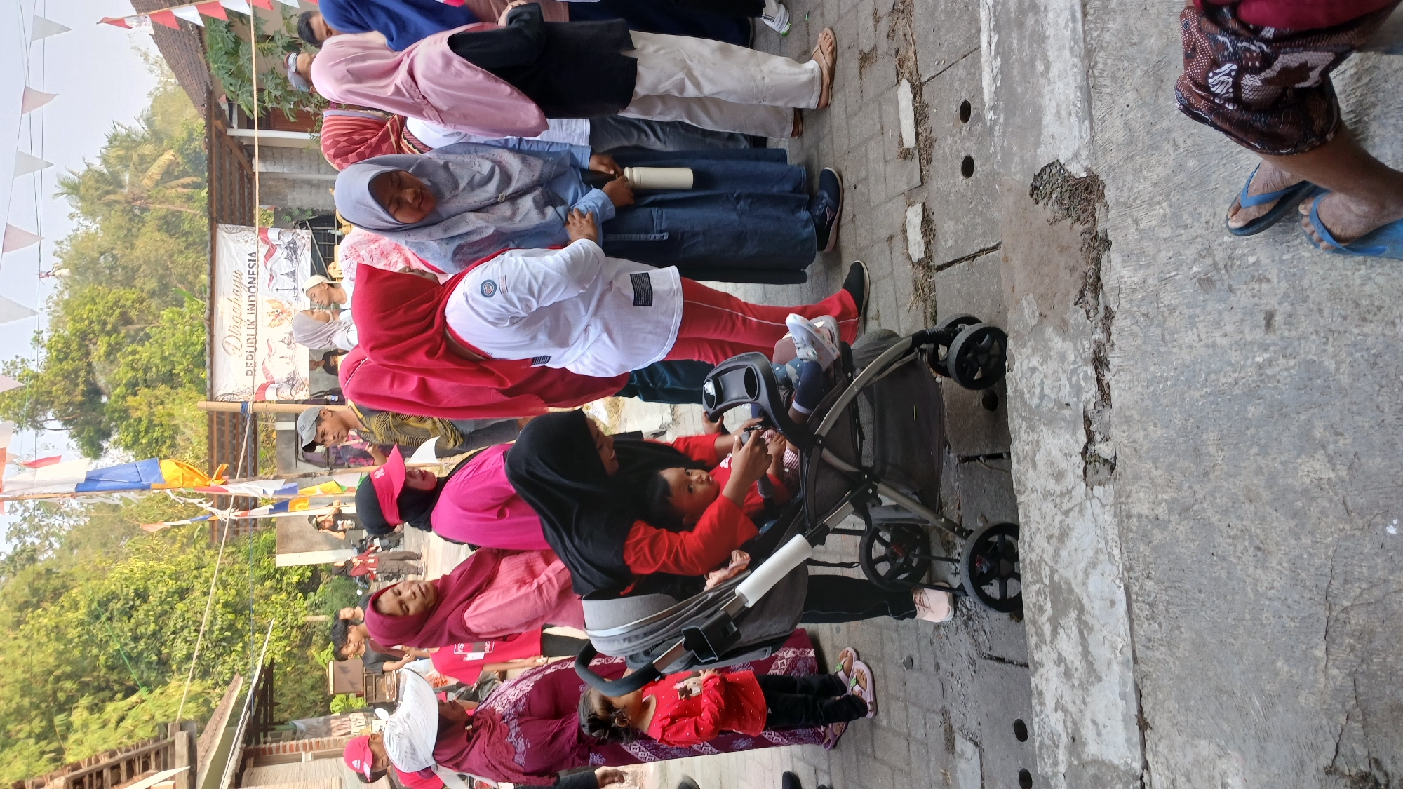 A group of people are gathered outdoors, possibly at a festival or event in Mendut, Indonesia. In the foreground, a woman in a black hijab and red shirt pushes a baby in a stroller. The baby, also in a red shirt, is looking at the camera. To the left, a young girl in a red shirt stands next to the stroller. In the background, several other people, mostly women wearing hijabs in various colors, are standing and talking. Some are wearing traditional Indonesian clothing. Flags and banners are visible, indicating a celebration or public gathering. The environment appears to be a street or pathway with concrete flooring and some greenery.