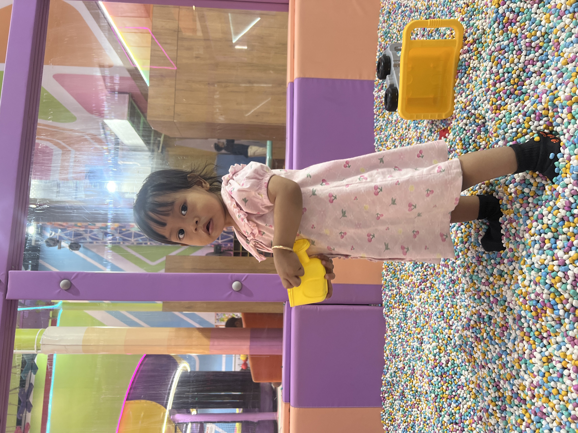 A young Southeast Asian girl, with dark hair and brown eyes, stands in a ball pit at an indoor playground. She is wearing a light pink dress with a cherry pattern and black socks. In her right hand, she holds a yellow toy watering can. To her right, a yellow toy dump truck is partially submerged in the colorful balls. The ball pit is surrounded by padded walls in shades of purple and orange. In the background, a colorful, multi-level play structure is visible through a clear partition. The overall environment suggests a children's entertainment facility.